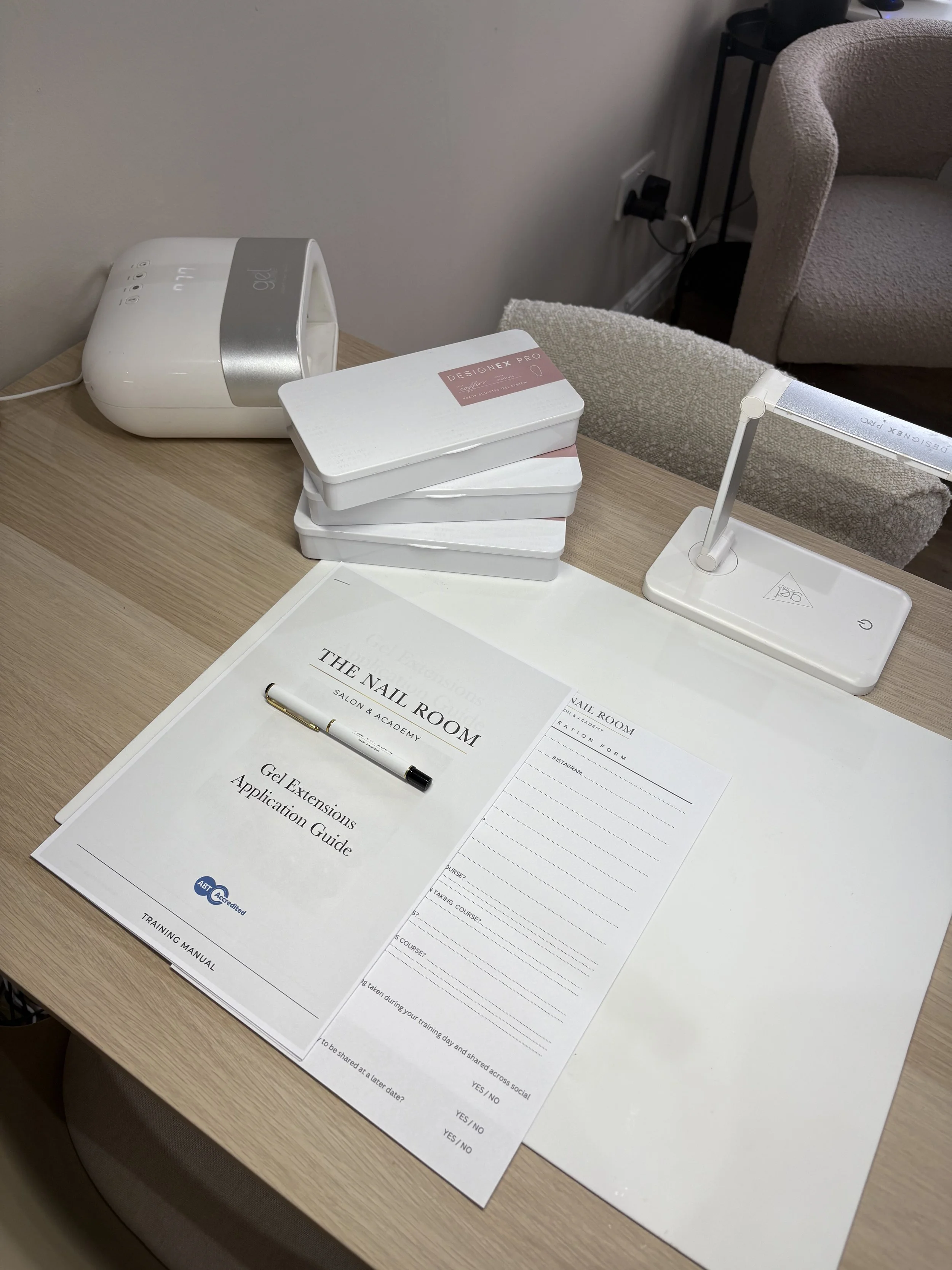 A desk with a nail salon training manual, application guide, a pen, a clipboard, several boxes of nail gel, a UV light, a desk lamp, and a chair in the background.