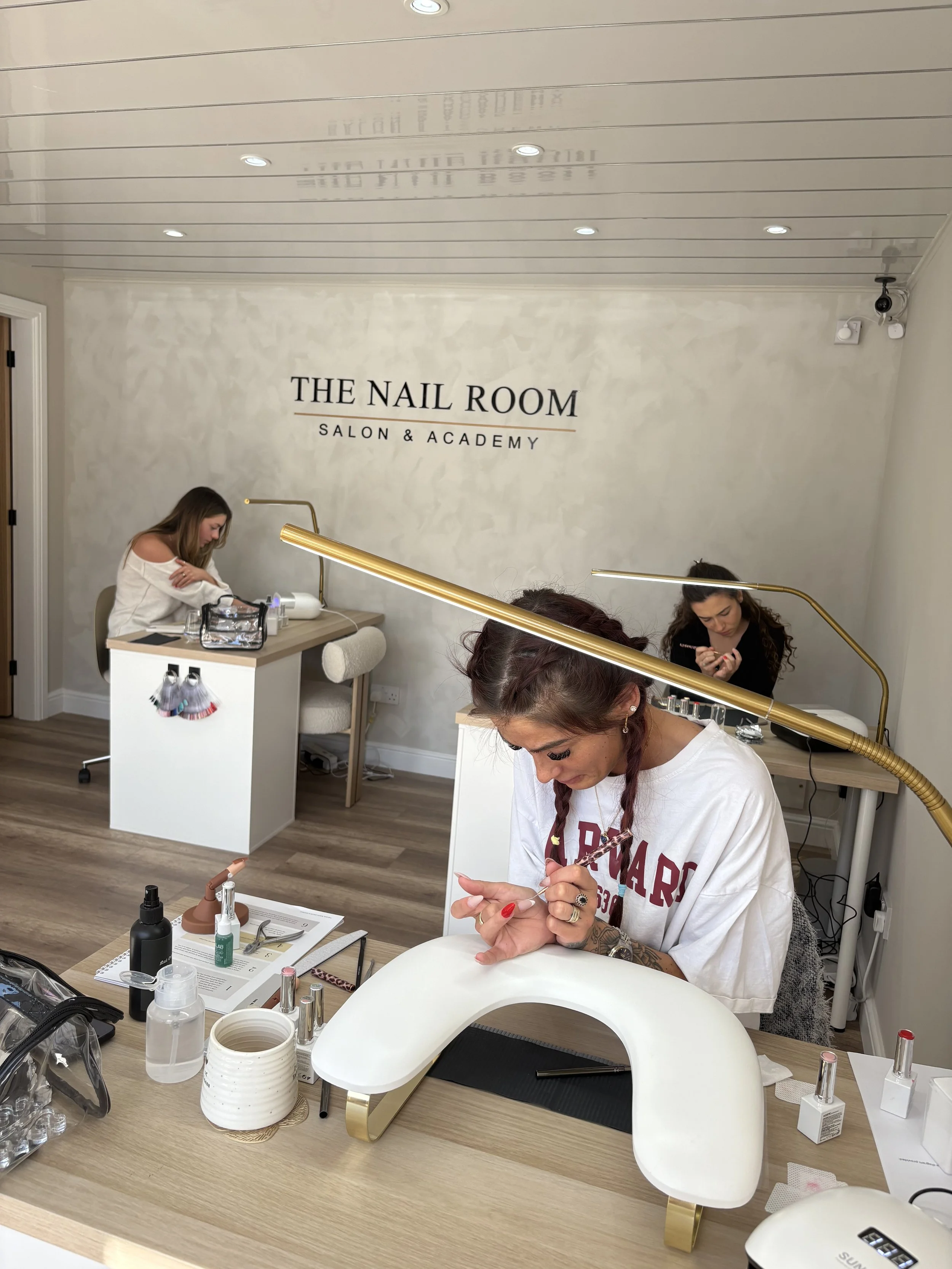 Three women working on nail art at a salon called 'The Nail Room Salon & Academy' with various nail tools and supplies on the table.
