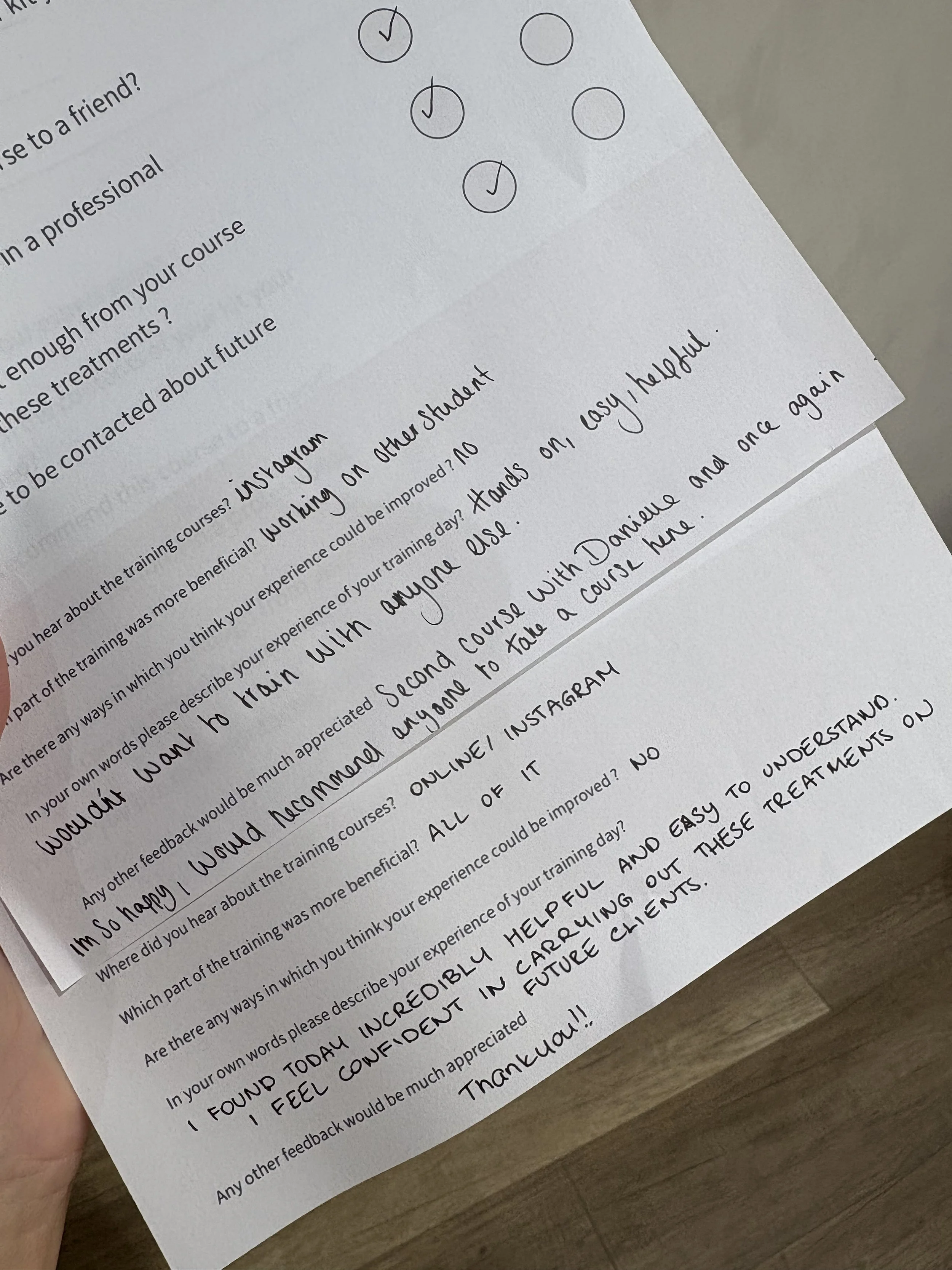 A handwritten feedback form on paper with checkmarks in circles at the top, and handwritten responses about training experience, including positive comments about online training and helpfulness, with a background of a wooden surface.