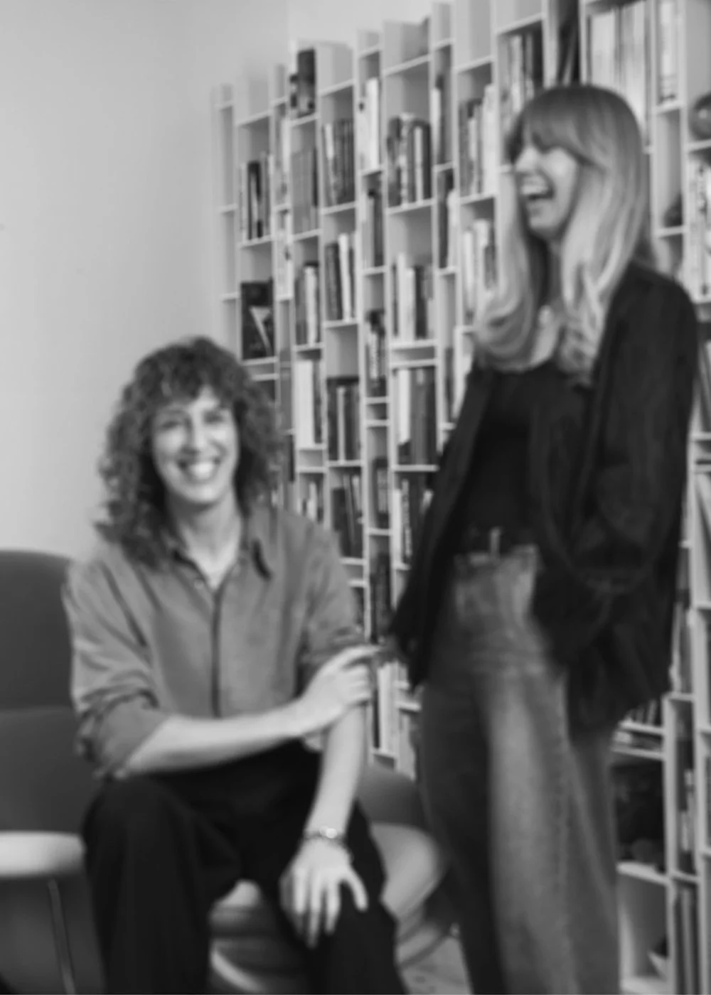Two women having a conversation in a library, one sitting and the other standing, both smiling.