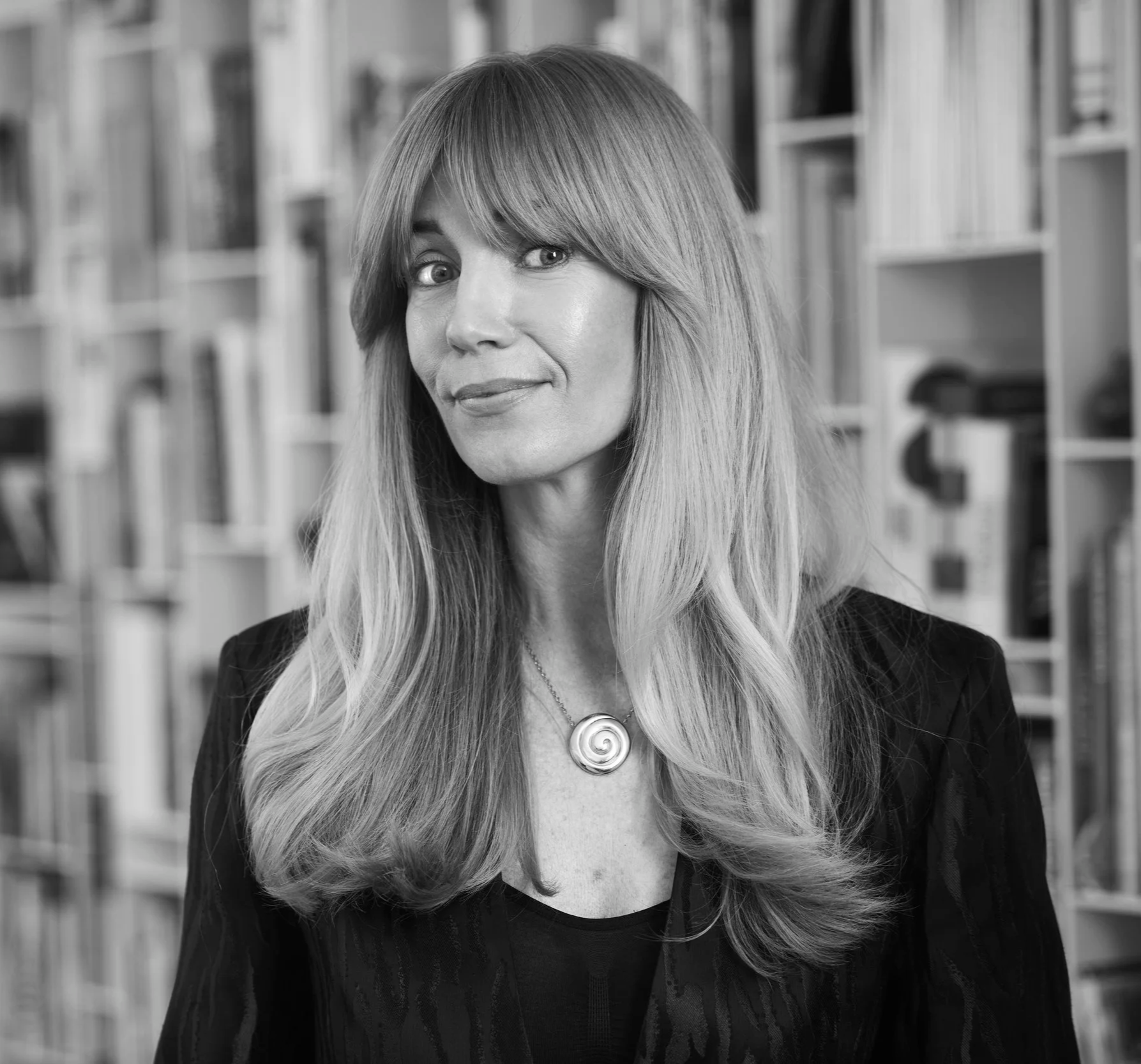 Black and white photo of a woman with long wavy hair, smiling slightly, wearing a necklace with a spiral pendant and a dark top, standing in front of bookshelves.