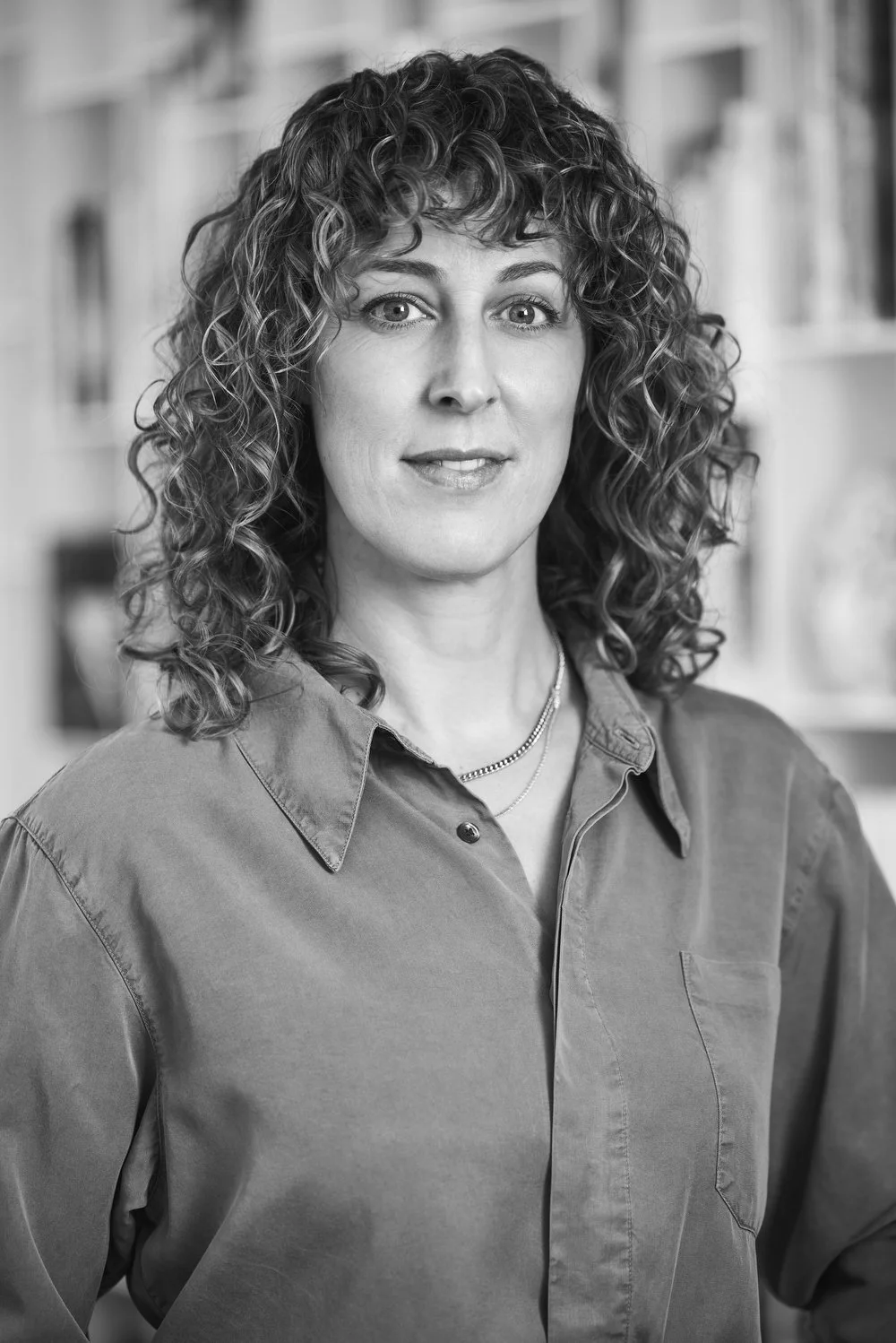 Black and white photo of a woman with curly hair wearing a collared shirt and layered necklaces, standing in front of shelves.
