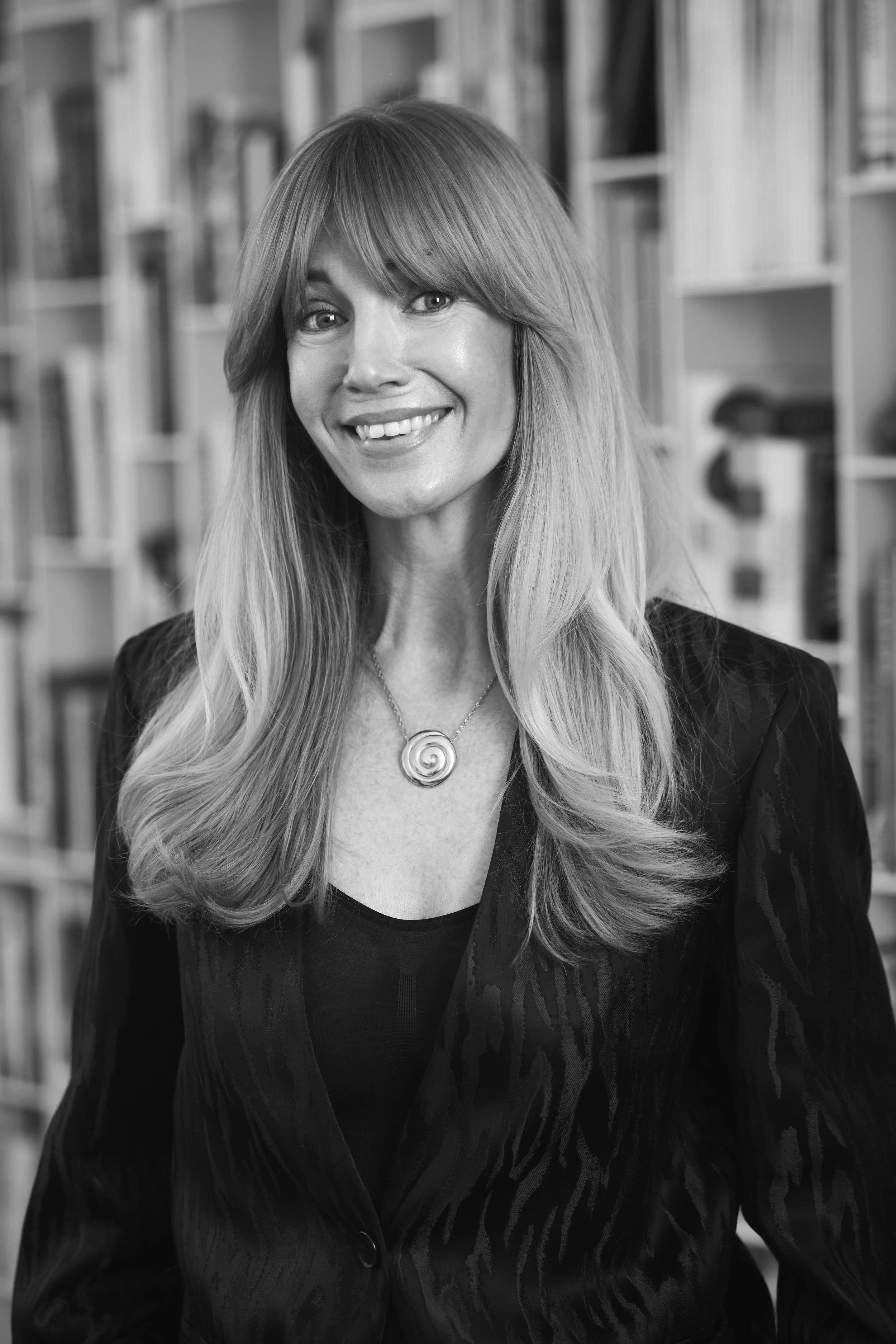 Black and white portrait of a woman with long, wavy hair, smiling, wearing a dark blazer and a necklace with a spiral pendant, in front of a bookshelf.