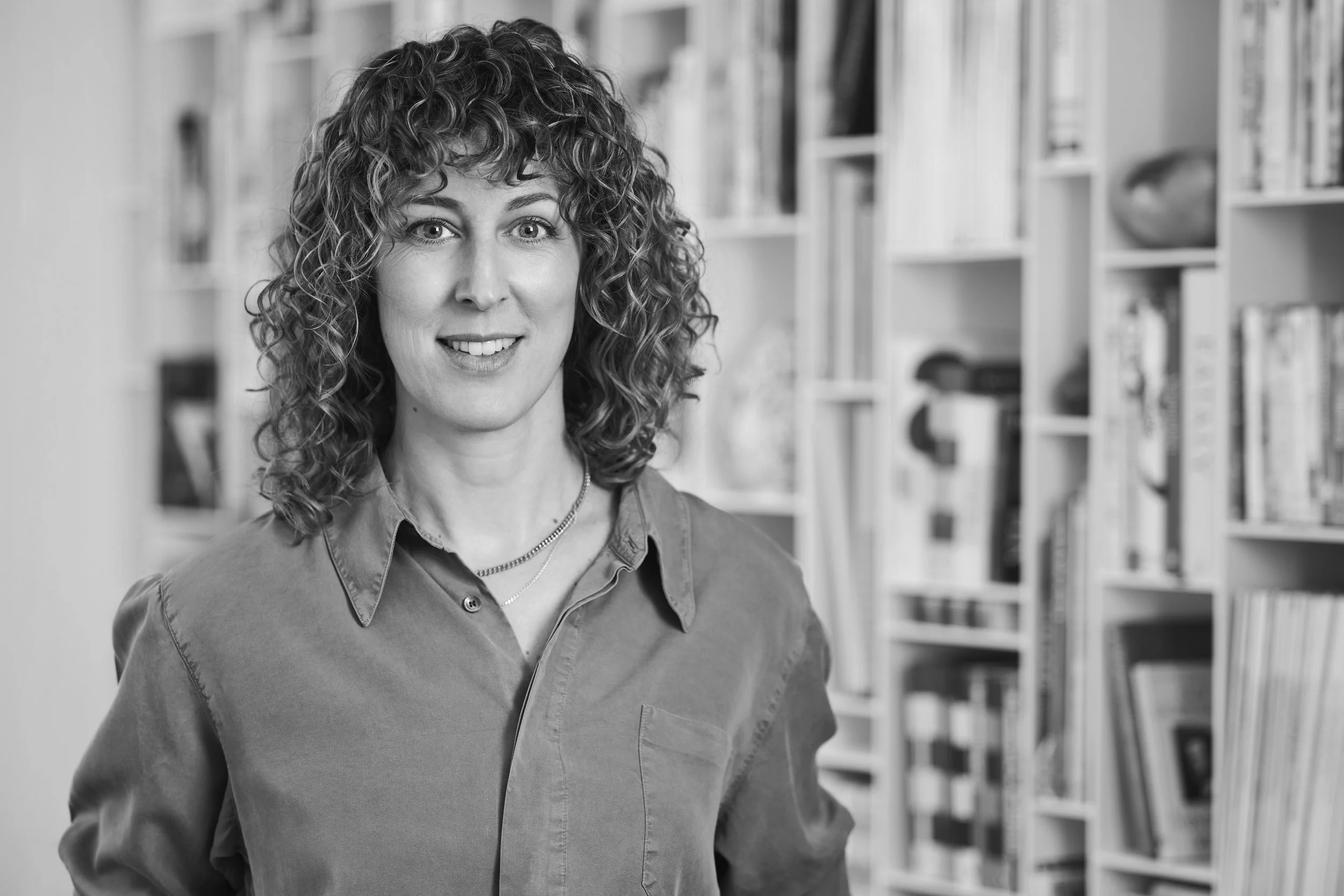 A woman with curly hair smiling in a library with shelves of books behind her.