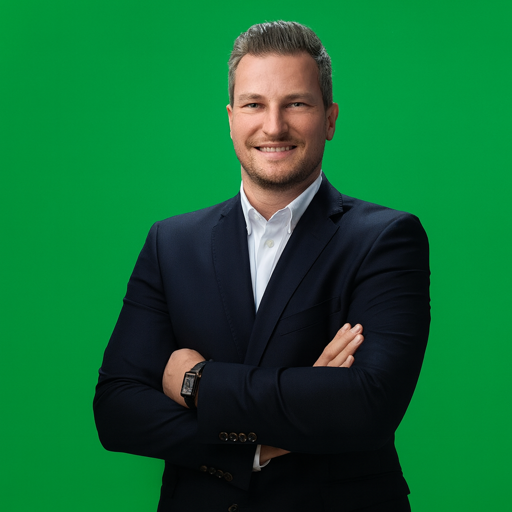 Ein Mann in einem dunklen Anzug und weißem Hemd steht vor einem grünen Hintergrund. Er lächelt und hat die Arme über Kreuz.