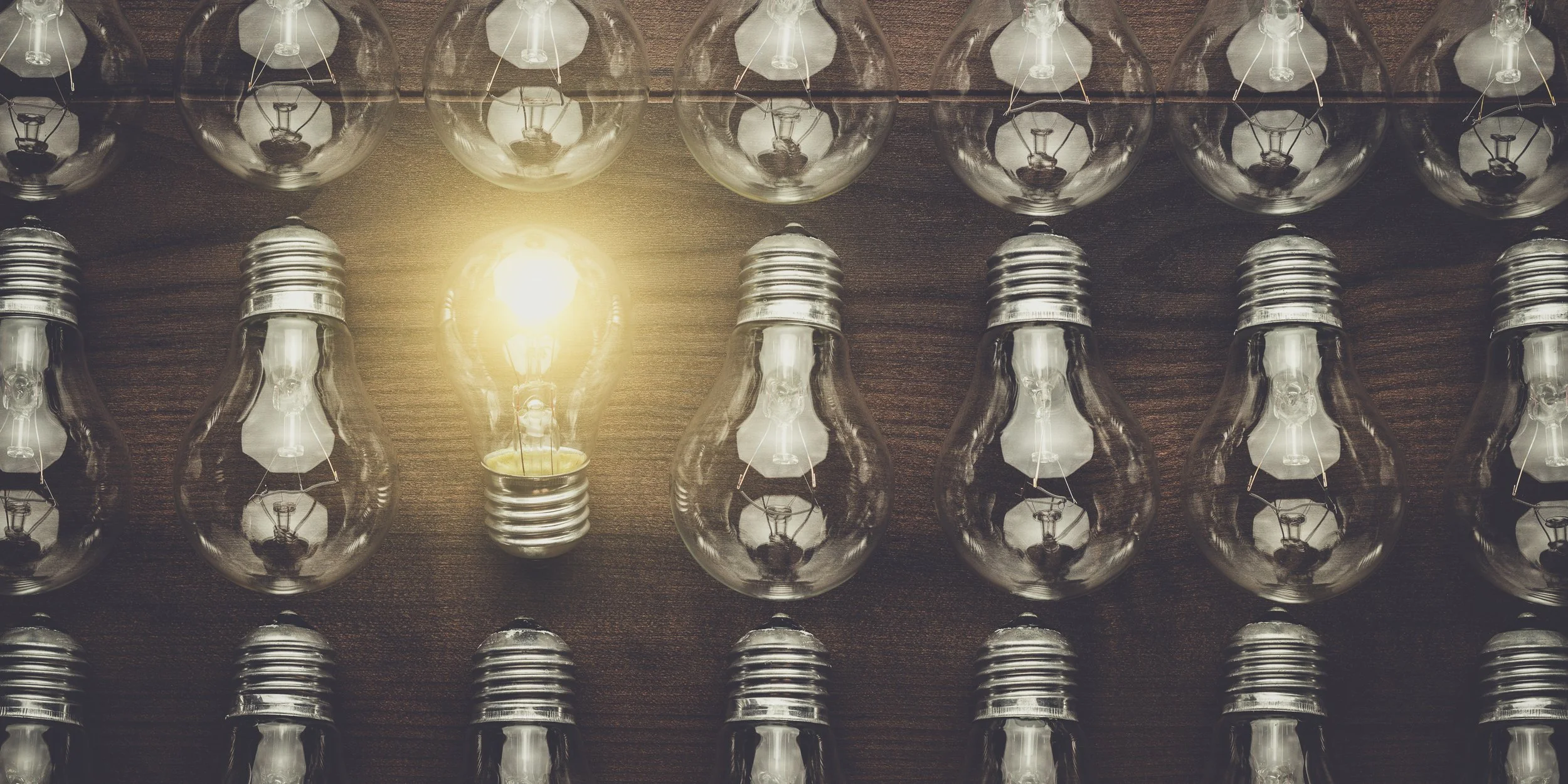 Mehrere Glühbirnen, eine davon leuchtet, auf einem Holzbrett. Die leuchtende Birne steht in der Mitte, die anderen sind ausgeschaltet.