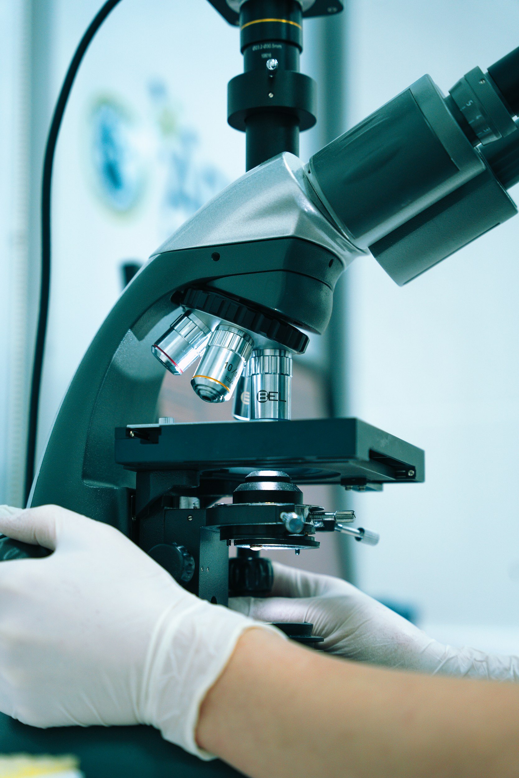 A gray laboratory microscope with glass lenses, held by someone wearing white gloves, in a laboratory setting.