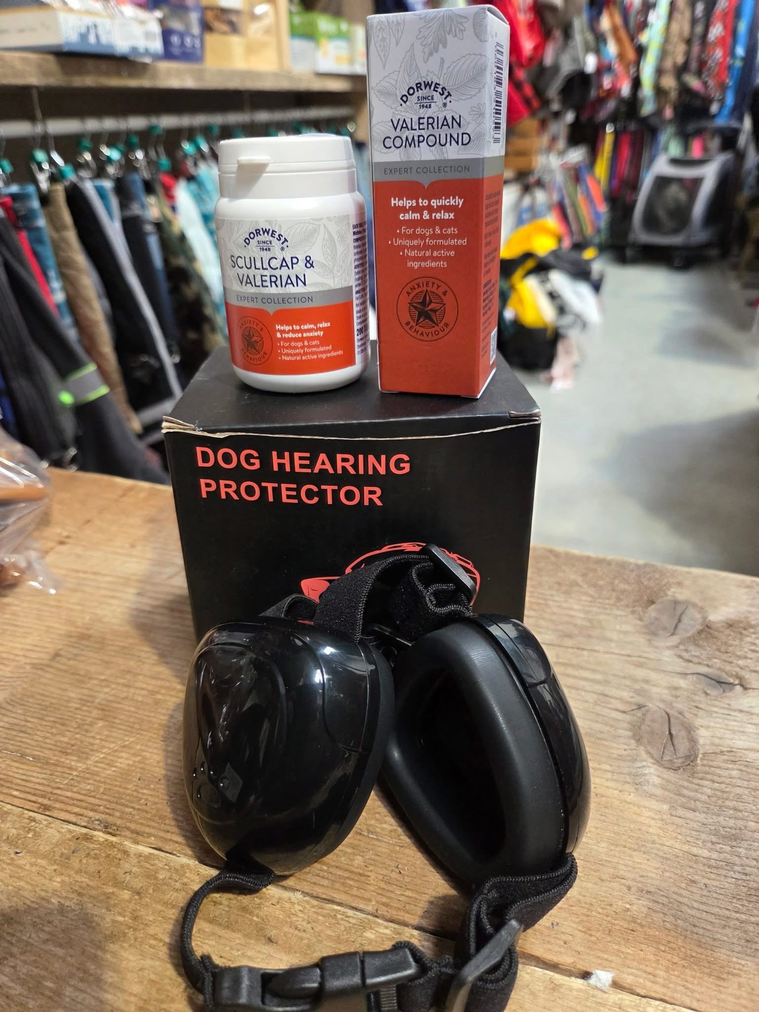 Dog hearing protector with black ear muffs and strap, dog calming supplements in a bottle and box, placed on top of a black box labeled "Dog Hearing Protector," in a pet or outdoor store.