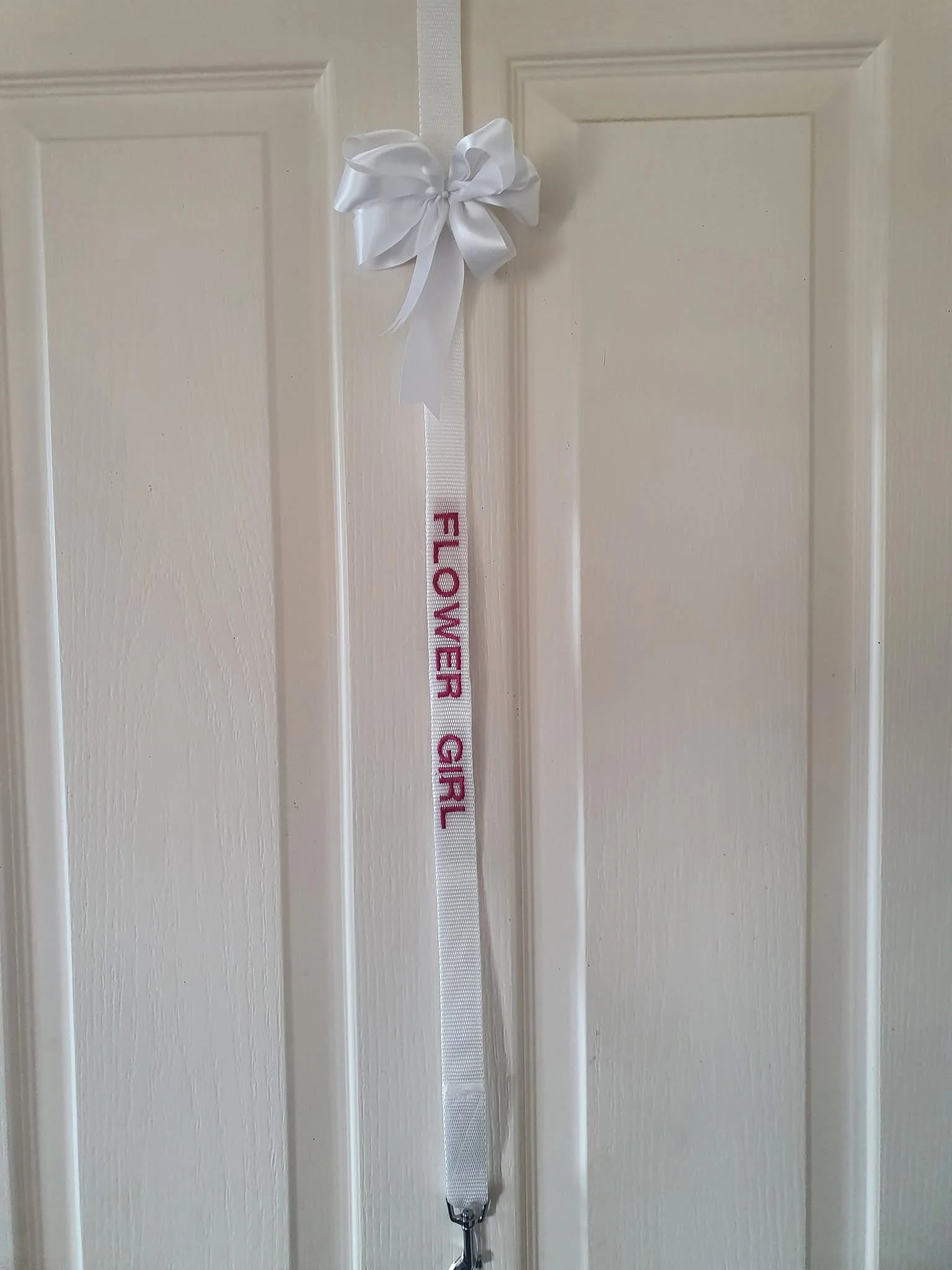 A white door with a decorative white ribbon bow at the top. Hanging from the door is a lanyard with the words "FLOWER GIRL" embroidered in pink.