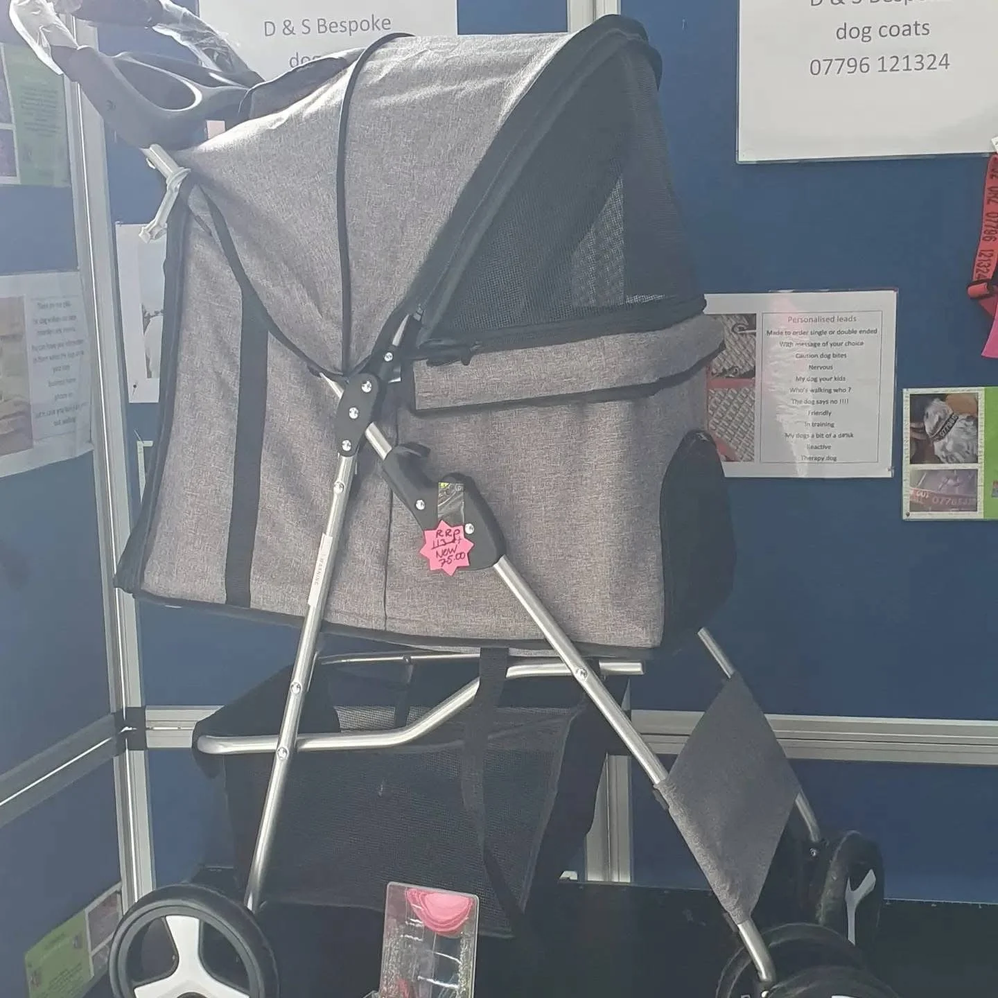 Gray baby stroller with black mesh side panels and attached footrest, displayed at a store on a black surface with a blue background and advertisements nearby.