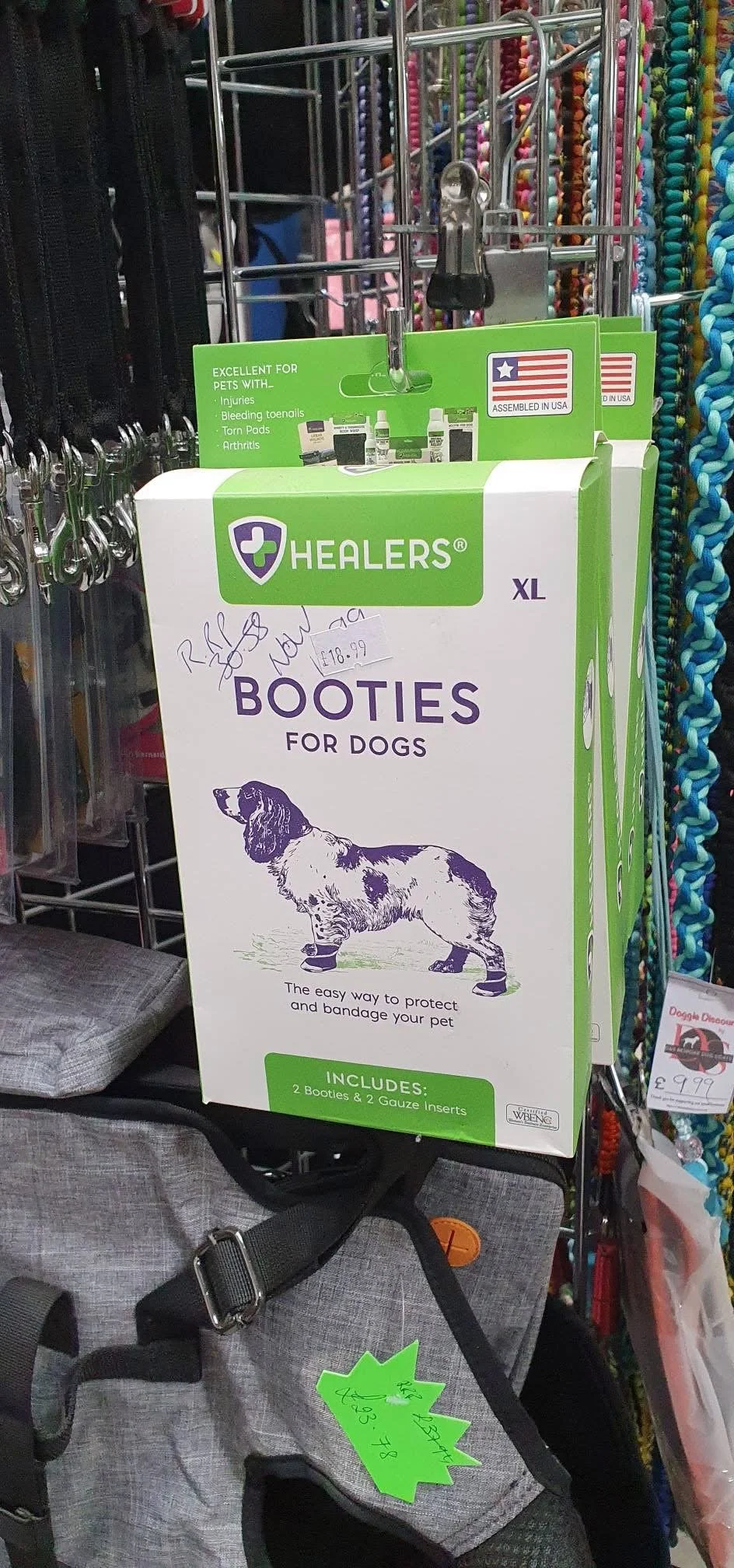 Box of dog booties for dogs with illustrations of a dog on the packaging, placed on a gray dog harness or bag display in a pet store, with various pet accessories visible in the background.