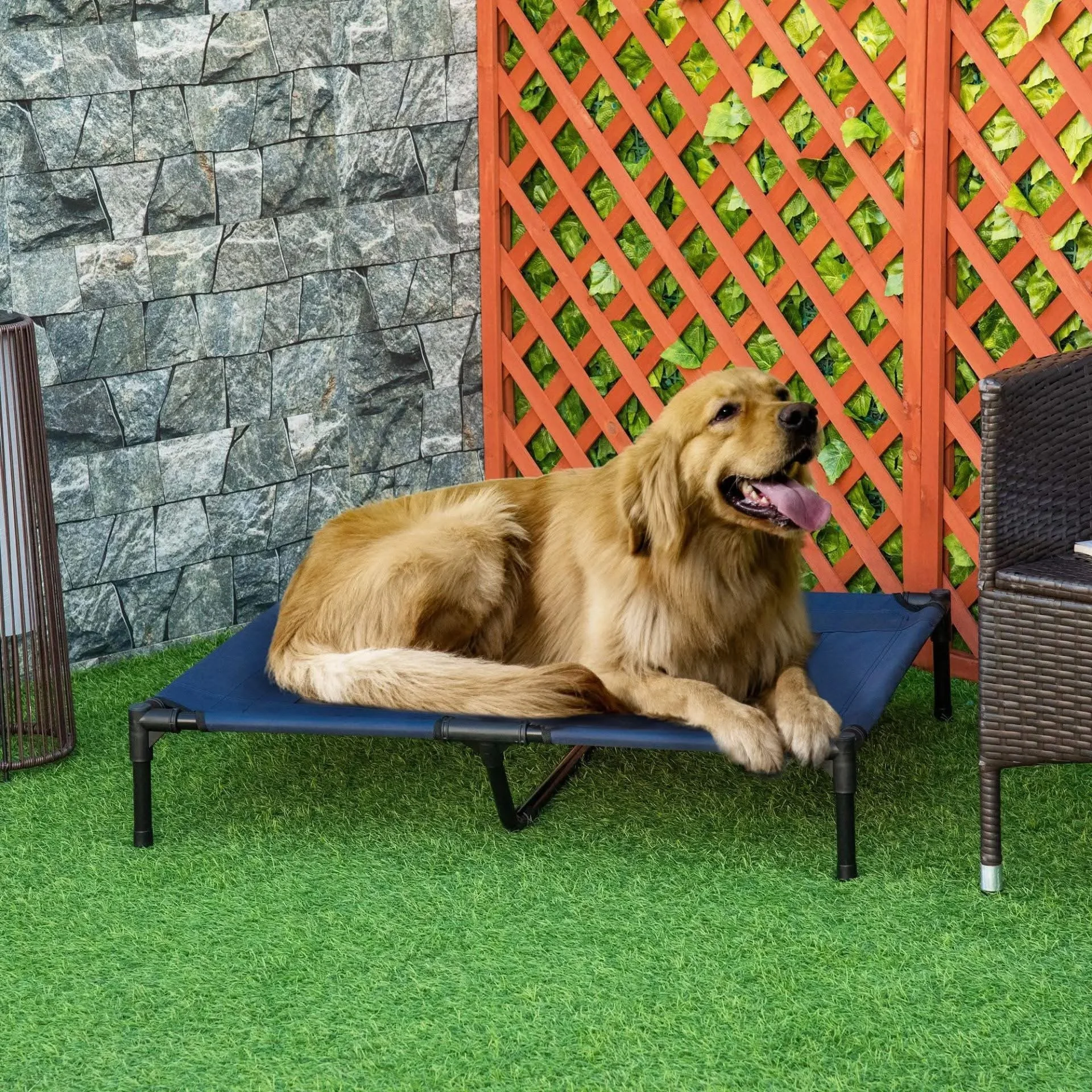 Golden retriever laying on a raised outdoor pet cot, panting with tongue out, in a backyard with green artificial grass, stone wall, and a red lattice fence with green foliage.