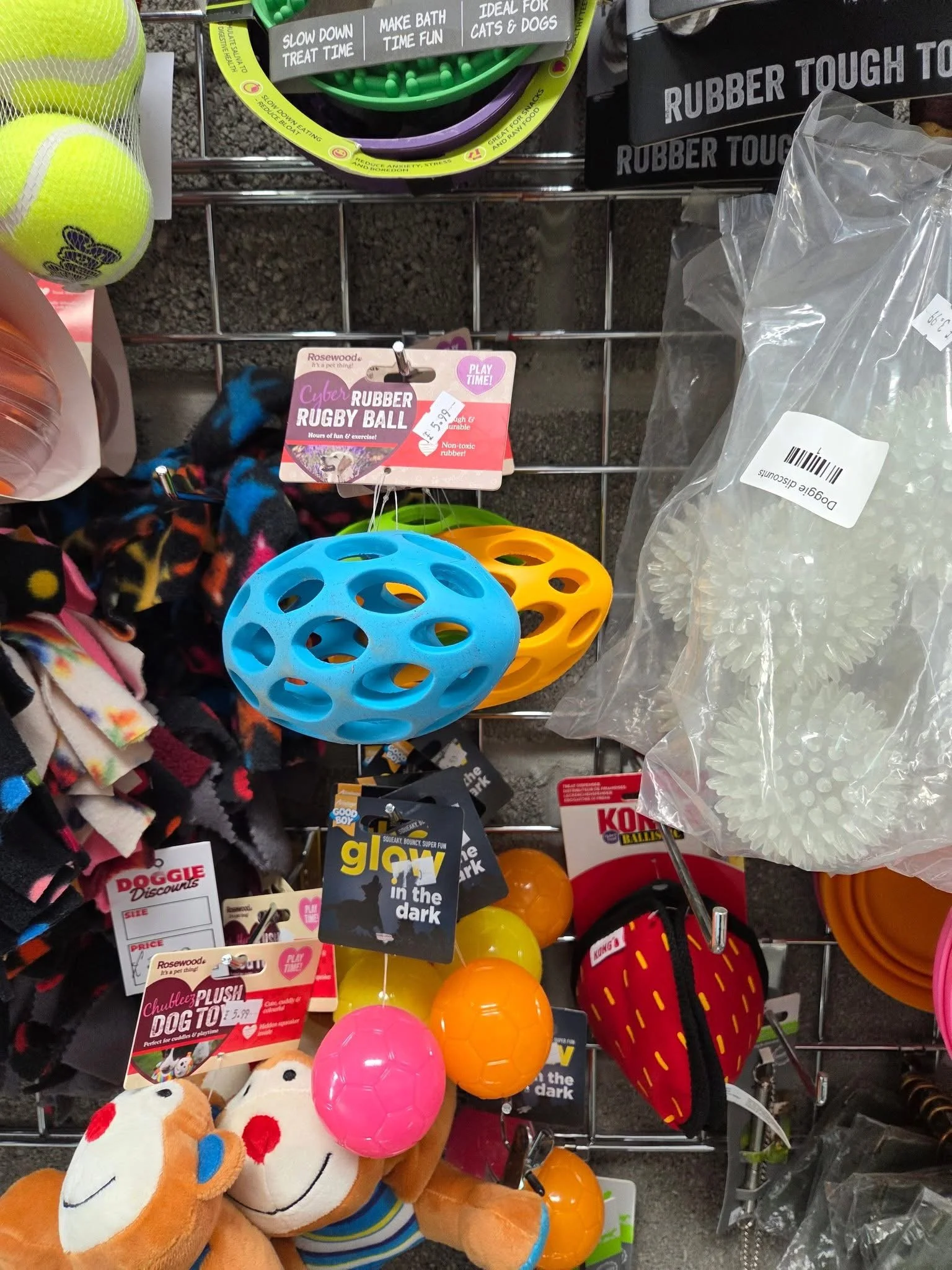 Colorful pet toys, including a blue rubber rugby ball and an orange ball, hanging on a display rack at a store.