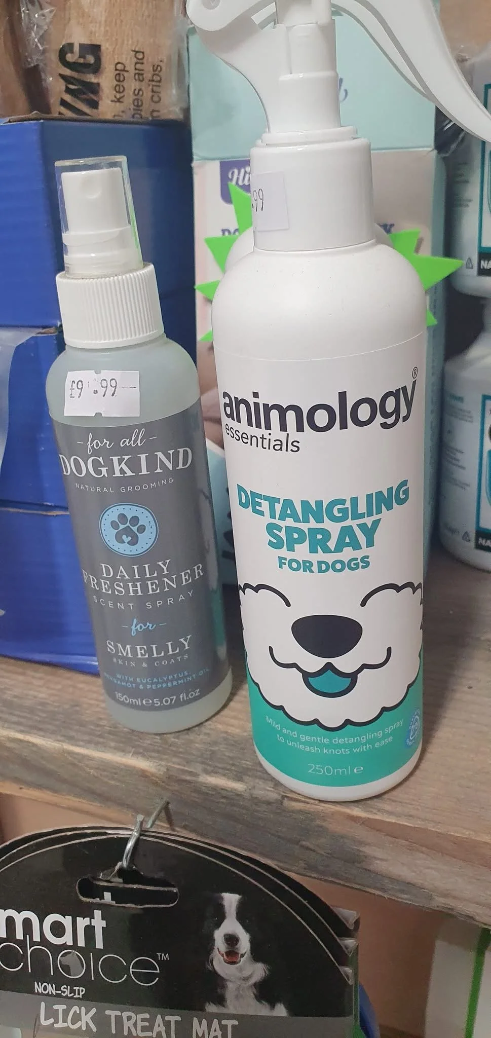 A white bottle of Animology Detangling Spray for Dogs and a smaller spray bottle of Dog Kind Daily Freshener for Smelly Skin and Coats are placed on a wooden shelf among pet grooming products.