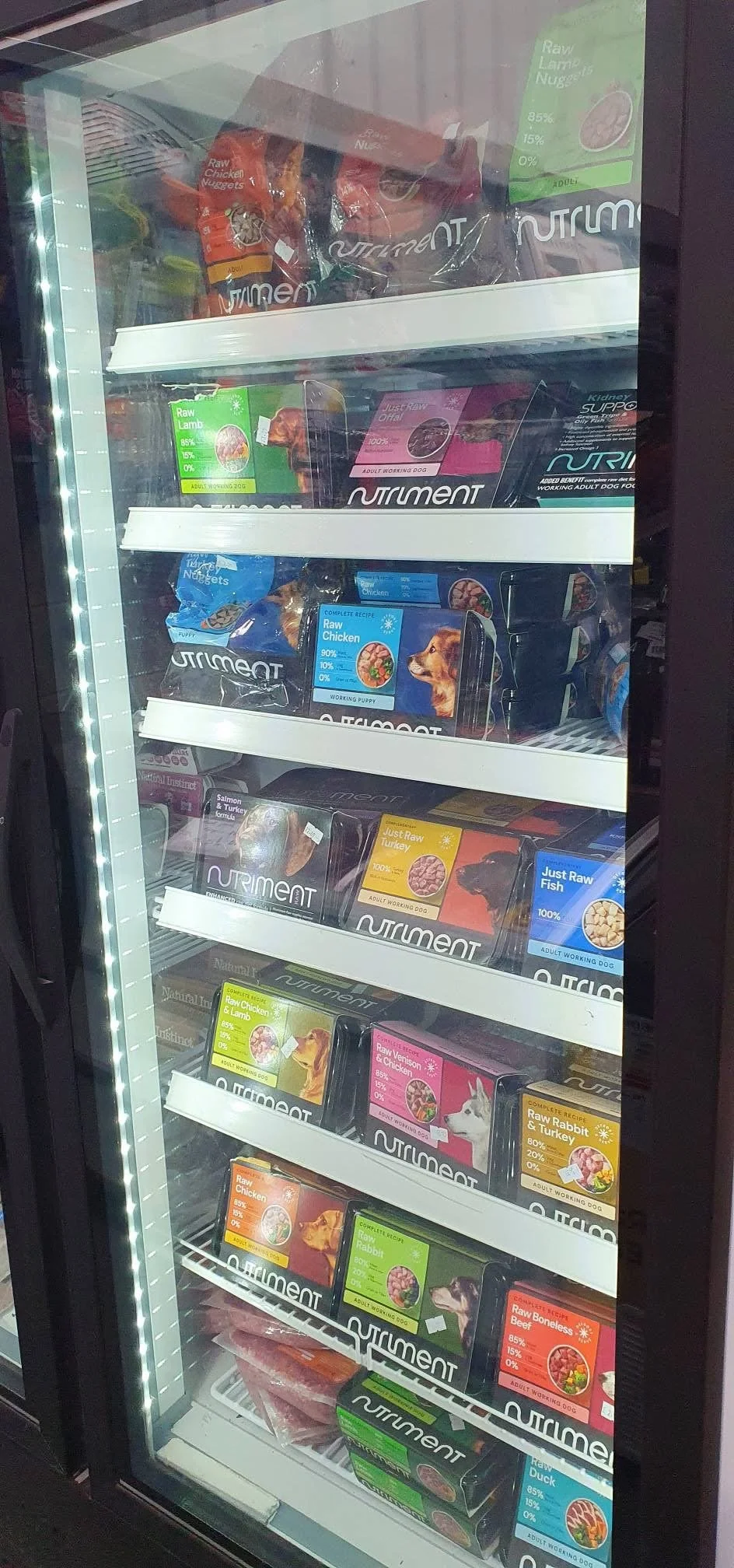 A meat freezer filled with various packages of raw pet food with colorful labels, including options like raw lamb, chicken, turkey, fish, rabbit, and venison, organized on shelves behind a glass door.