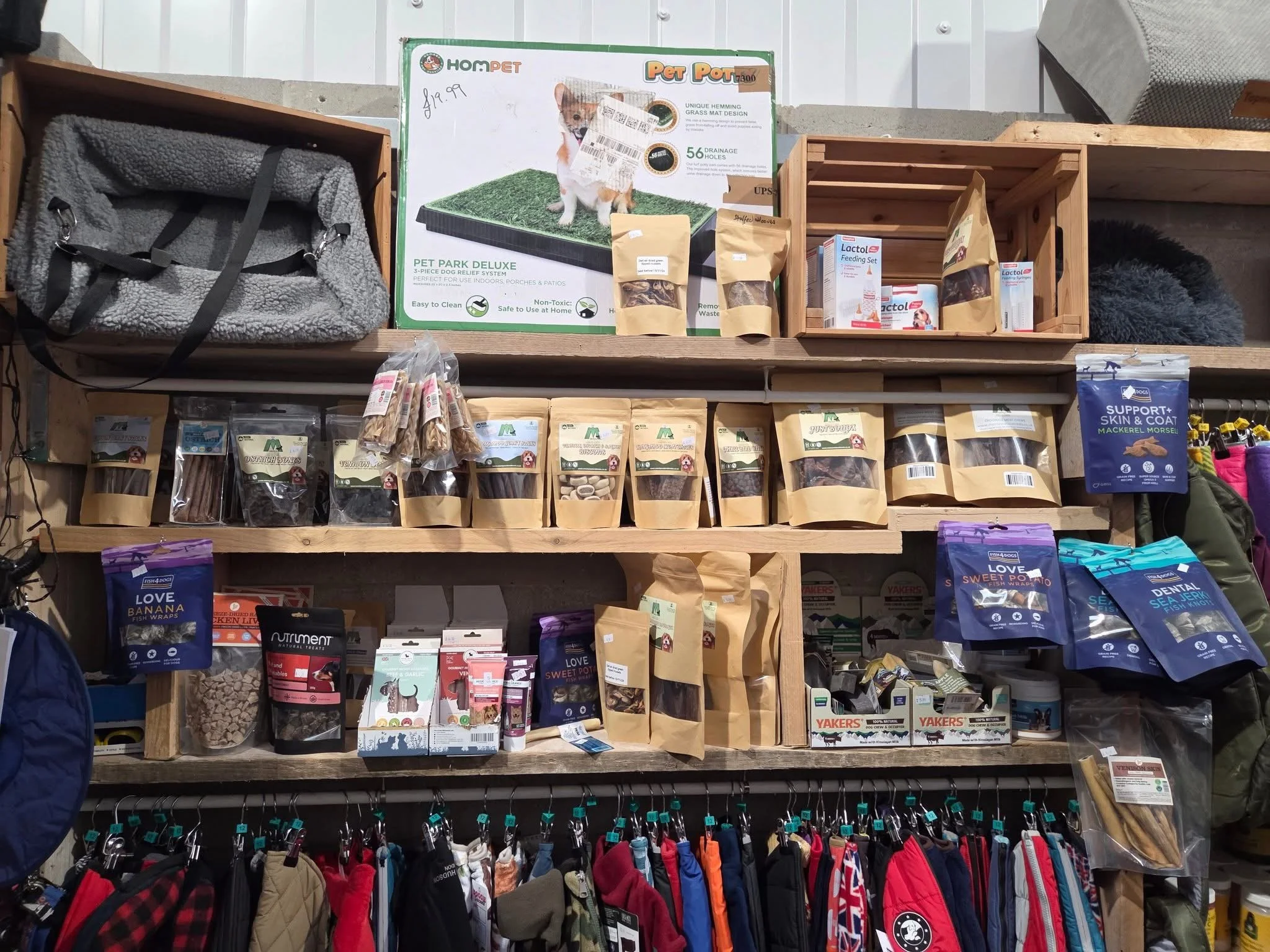 Shelves of various pet products, including bags of dog treats, pet toys, and grooming supplies, with clothing racks at the bottom displaying jackets and harnesses.