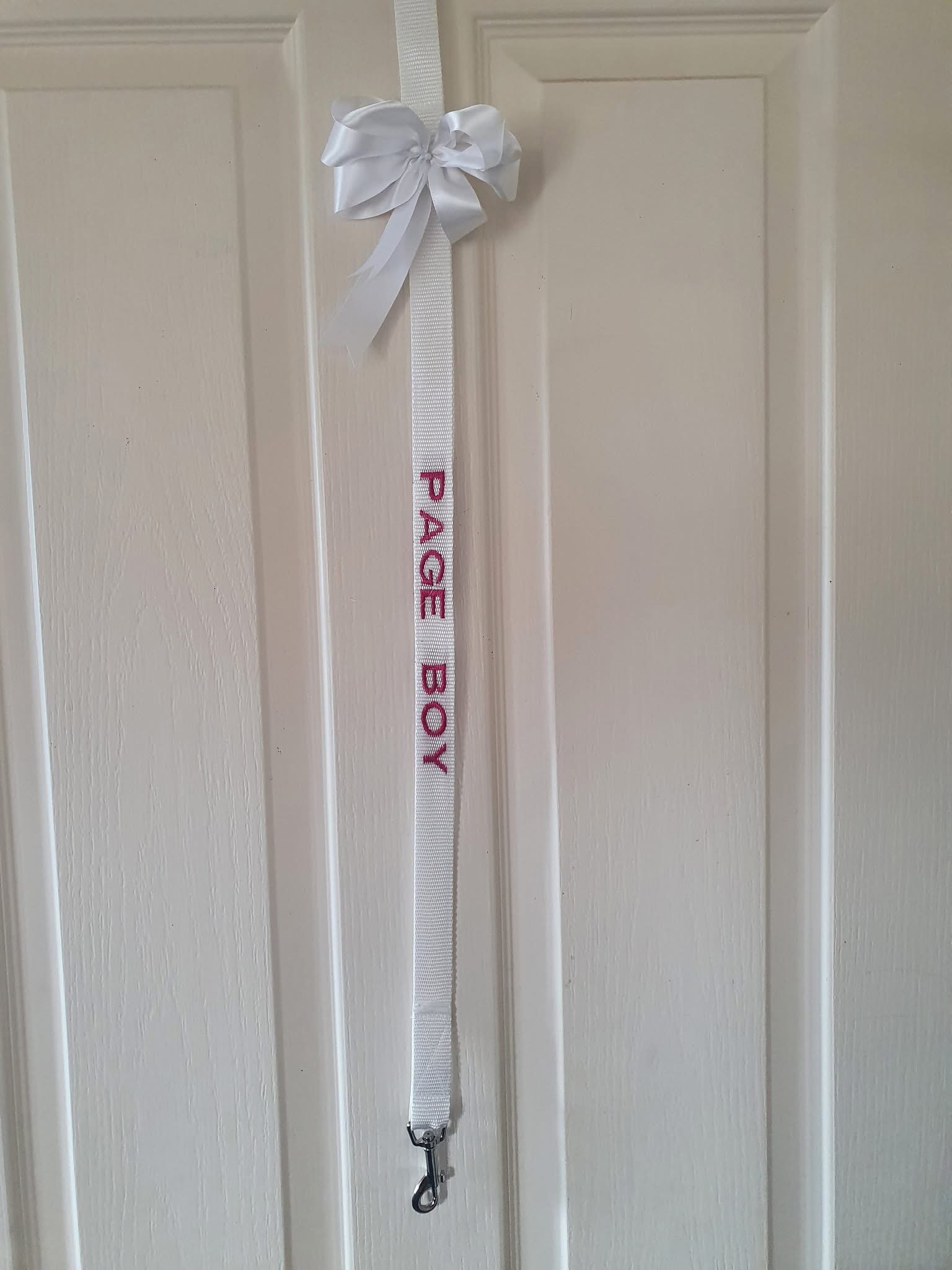 A white pet leash with red embroidery reading 'PEACE BOY' hangs on a white door, with a large white bow at the top of the leash.
