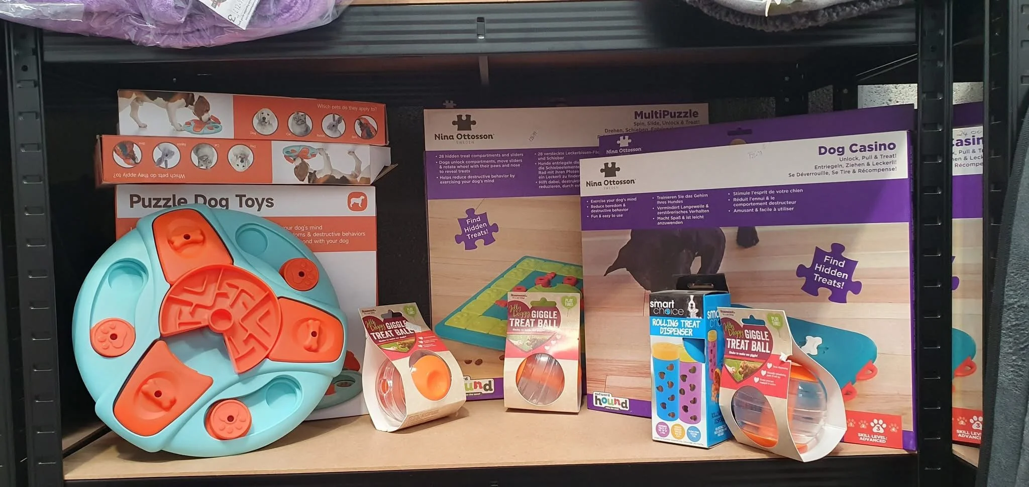 Shelf with various dog puzzle toys and treat dispensers, including a large orange and blue circular puzzle toy, a giggle treat ball, a rolling treat dispenser, and other pet puzzle items with packaging.