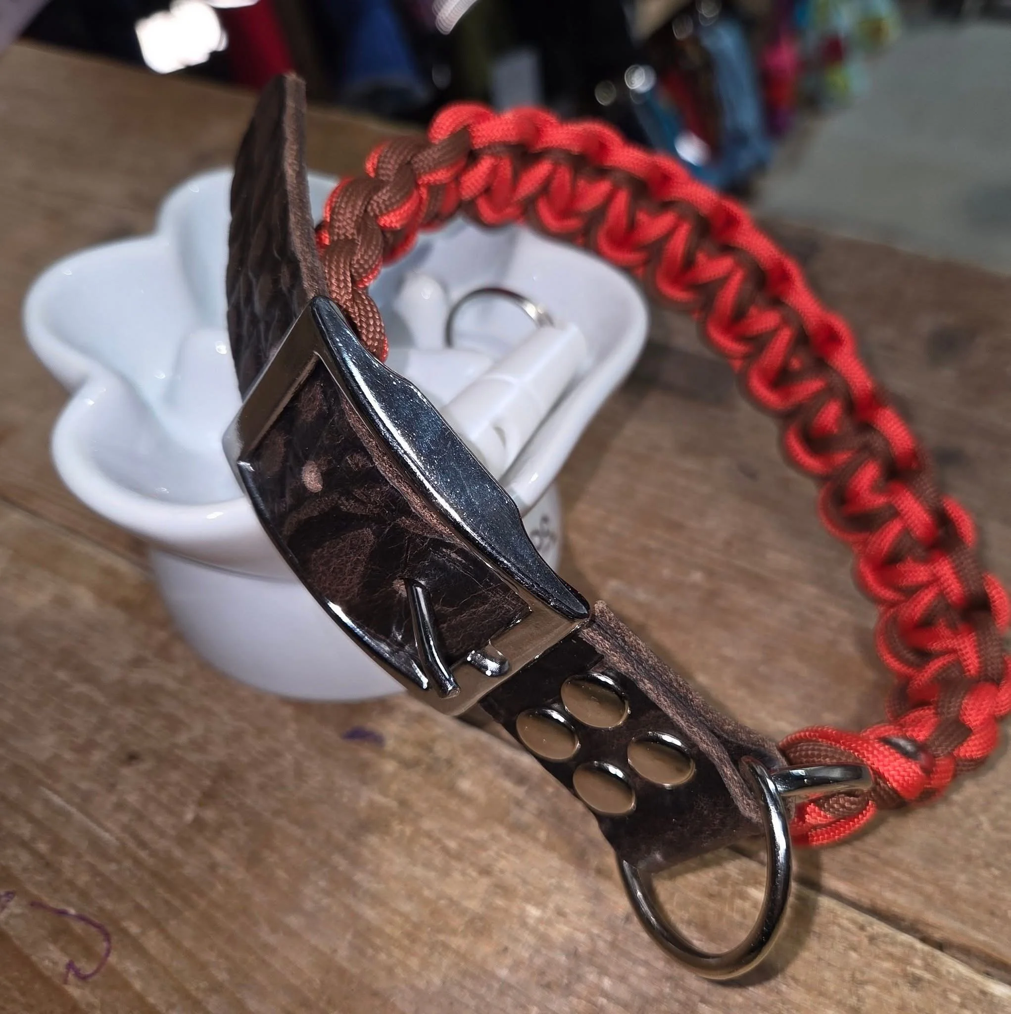 A leather dog collar with a metal buckle and red and brown paracord braided leash, placed on a wooden surface with a white ceramic container in the background.
