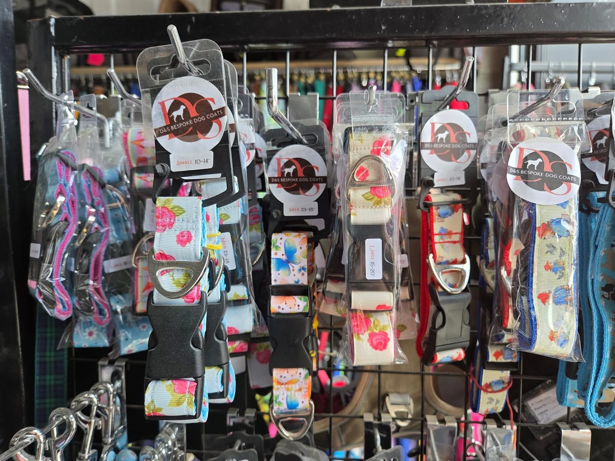 Display of colorful dog collars and harnesses hanging on a black metal grid, with tags indicating sizes and designs.