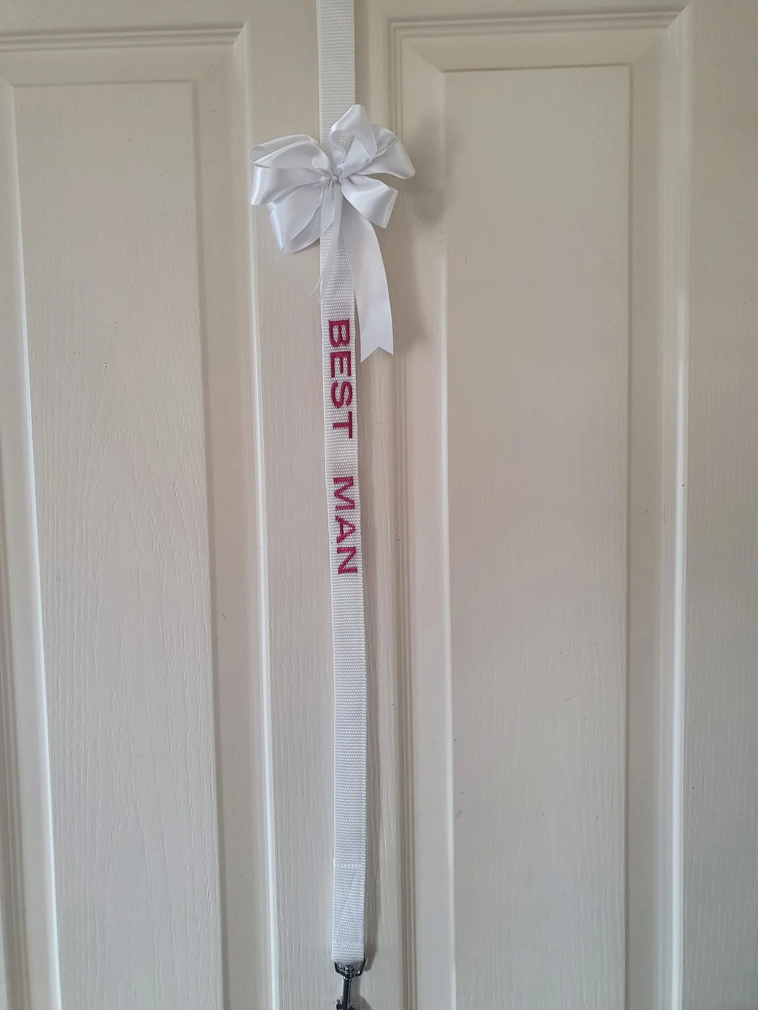 White door with a white ribbon and bow hanging from the top, attached to a white lanyard with the words 'BEST MAN' embroidered in pink.