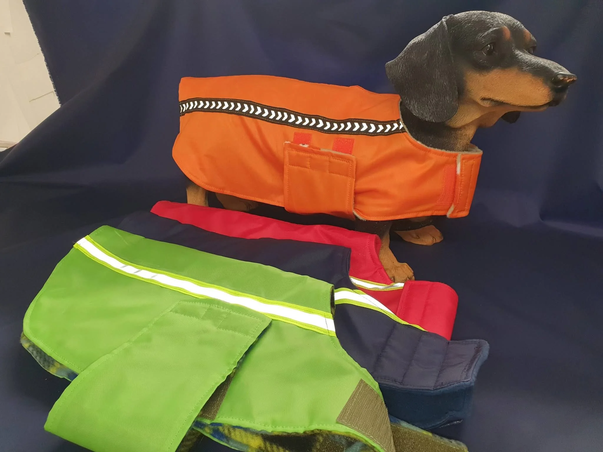 Dachshund dog wearing an orange safety vest with reflective strip, sitting on a dark surface. There are also two additional safety vests: one green with reflective strip and one red with black, lying on the surface.