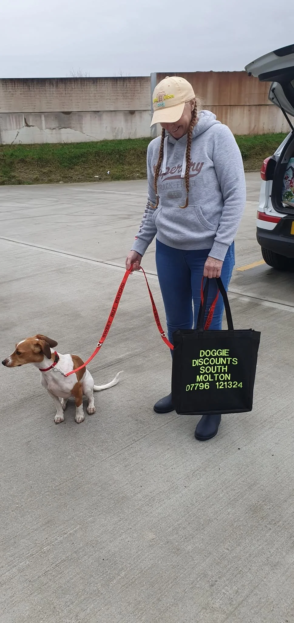 A woman with braids, wearing a beige cap with colorful text, a gray hoodie, and blue jeans, is holding a small dog on a leash. The dog is sitting on the street, and the woman holds a black bag with bright green text advertising a dog service.