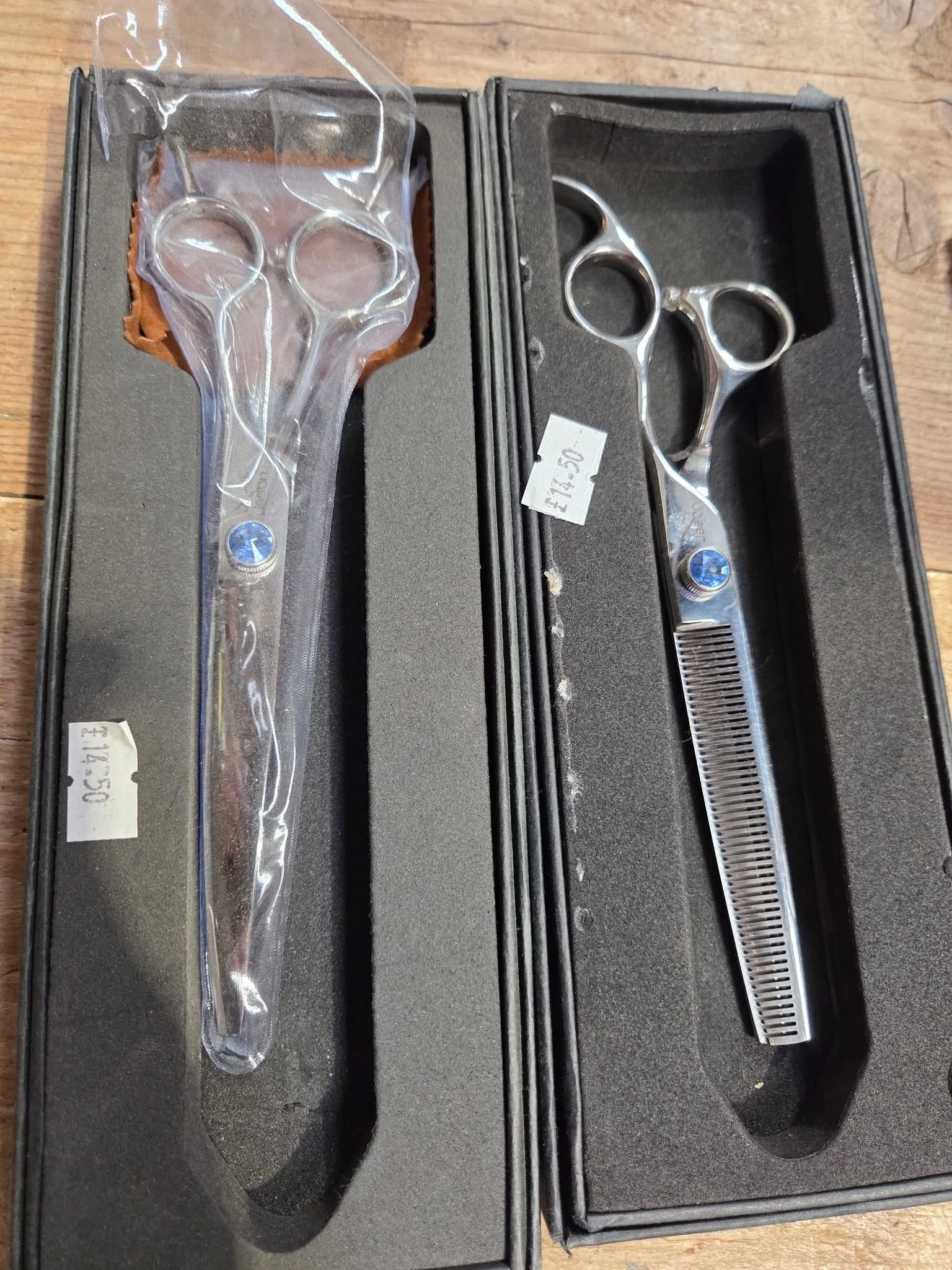 A set of hairdressing scissors in a foam-lined case, with a pair of thinning scissors in a plastic bag, displayed on a wooden surface.
