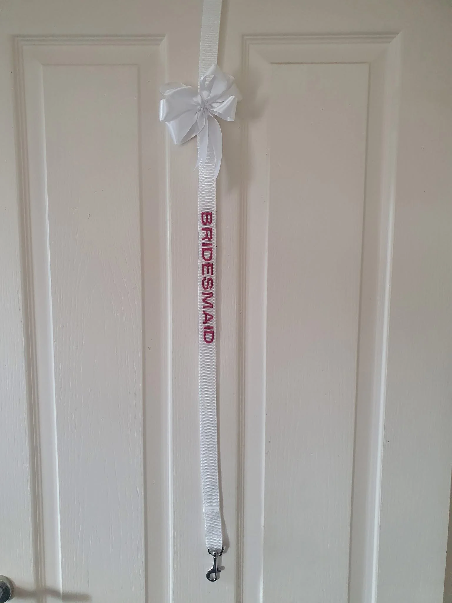 White wedding lanyard with the word 'BRIDESMAID' in pink, a white bow at the top, hanging on a door.