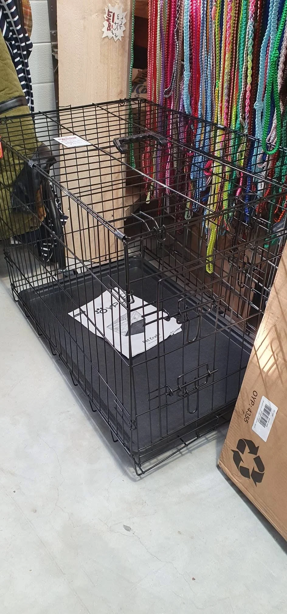Empty black metal pet crate in a store with colorful beaded necklaces hanging on a display behind it.