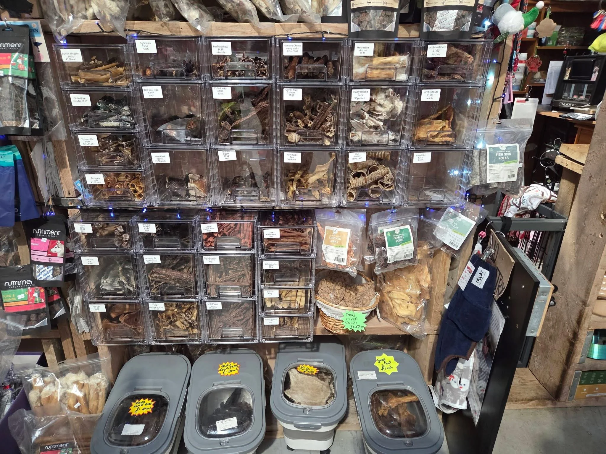 A display shelf with multiple clear plastic containers filled with various dried animal parts, bones, and tripe, labeled with their contents and prices. Below the shelf are additional containers with dried animal parts, some with hand-written price t