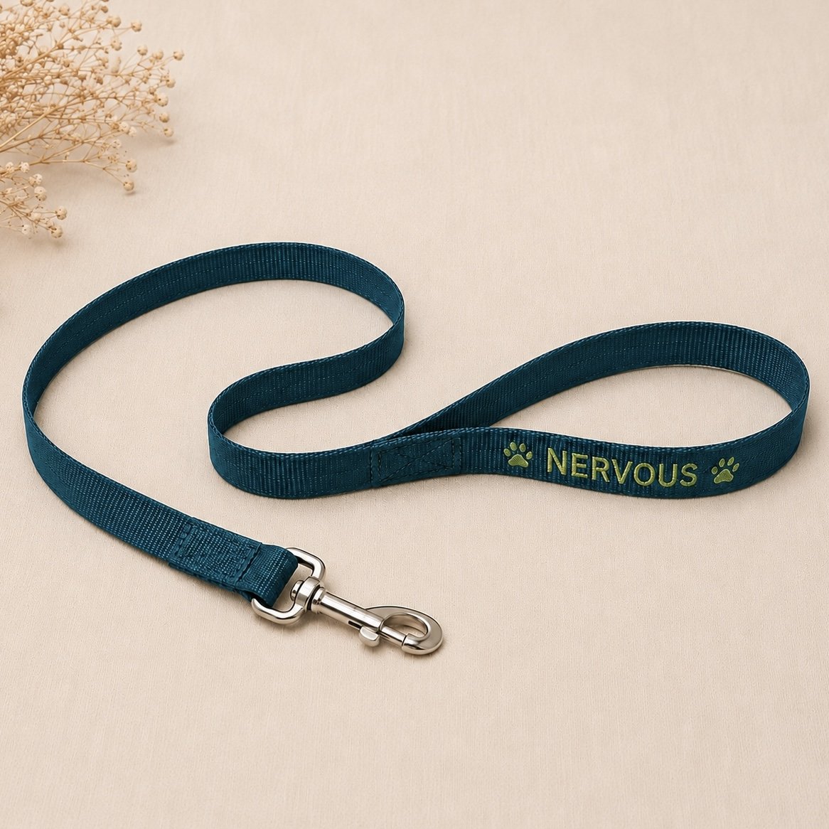 A blue pet leash with a metal clip and the word "Nervous" embroidered in yellow along with two paw prints, placed on a light-colored surface with a small bunch of dried flowers in the upper left corner.