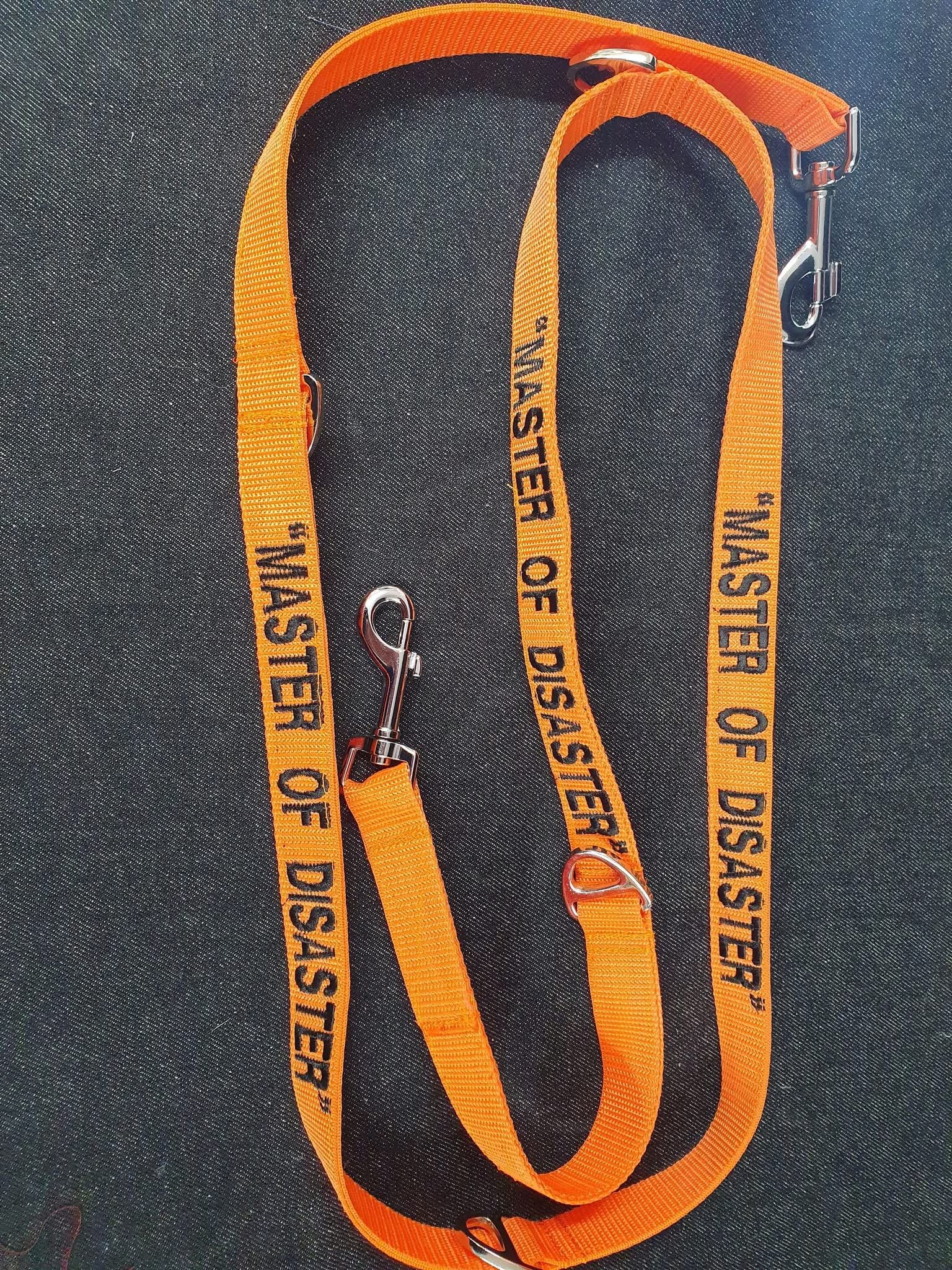 Orange safety lanyard with metal hooks, labeled 'Master of Disaster' repeated along its length, placed on a dark fabric background.