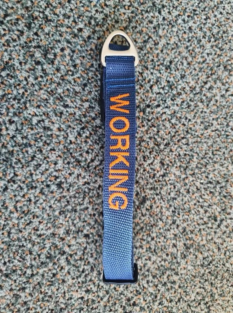A blue fabric belt with yellow embroidered text that reads 'WORKING', attached to a white and black metal clip, lying on a speckled gray carpet.