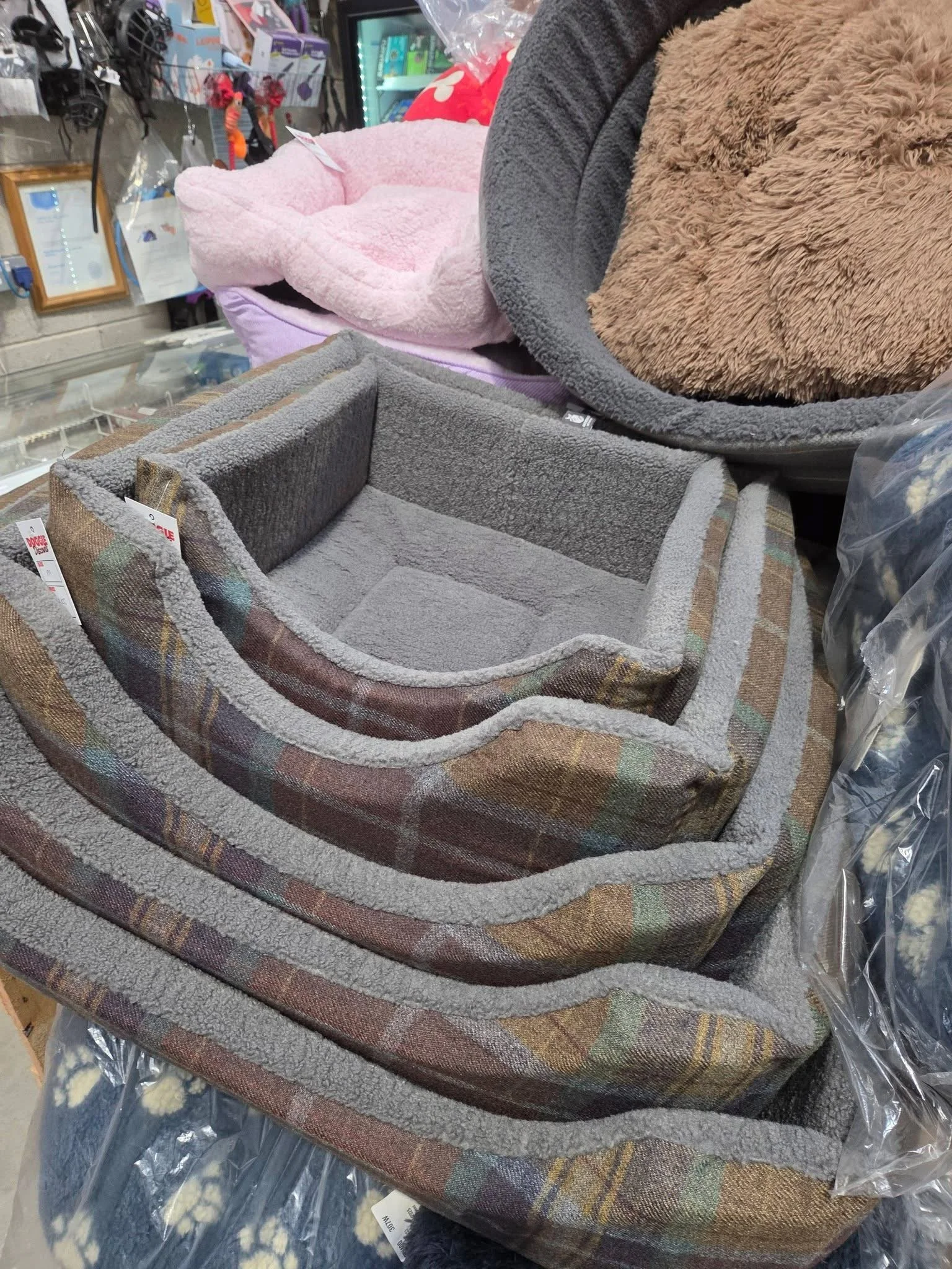 A display of cozy pet beds in a store, including a plaid fabric bed with a soft gray interior, and other plush beds in pink, brown, and gray in the background.