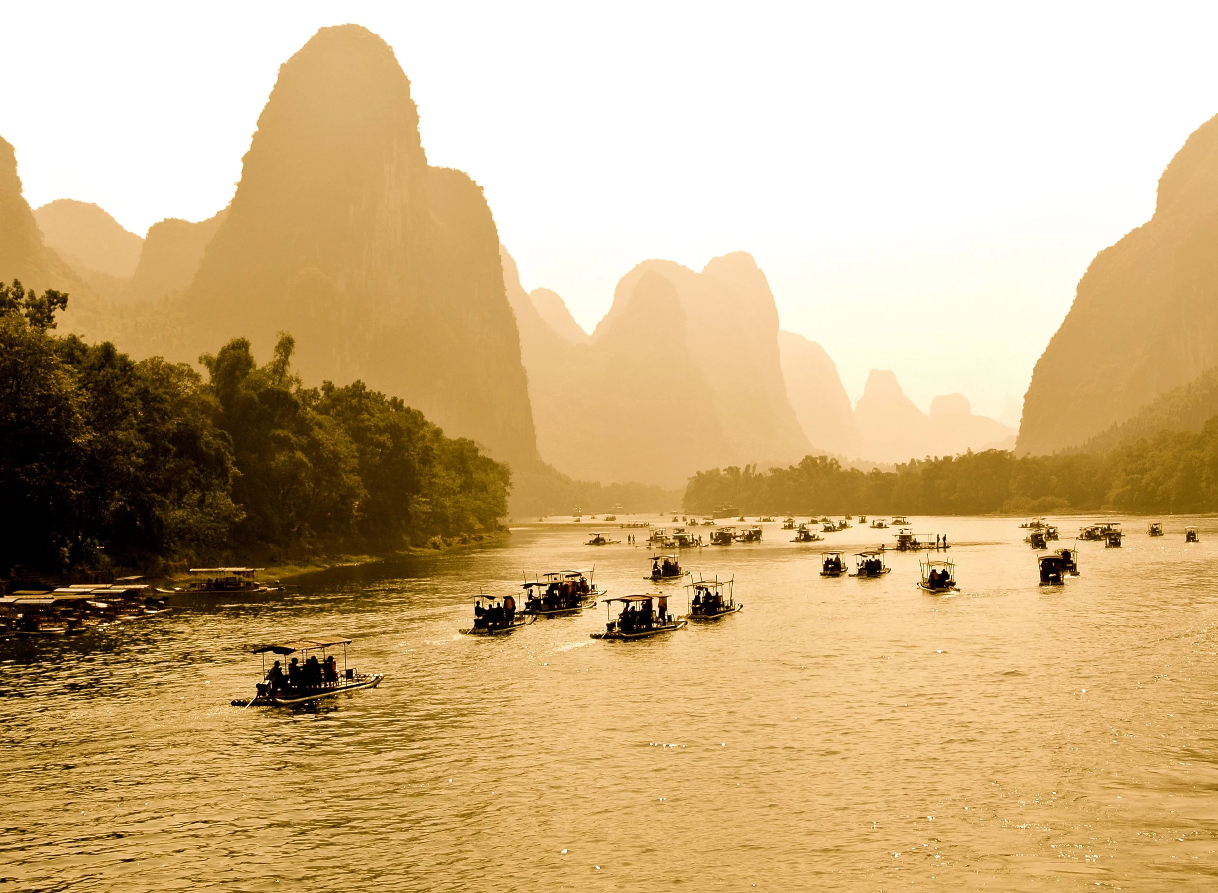 Boats floating on a river surrounded by lush green trees and tall limestone karst mountains under a hazy sky.
