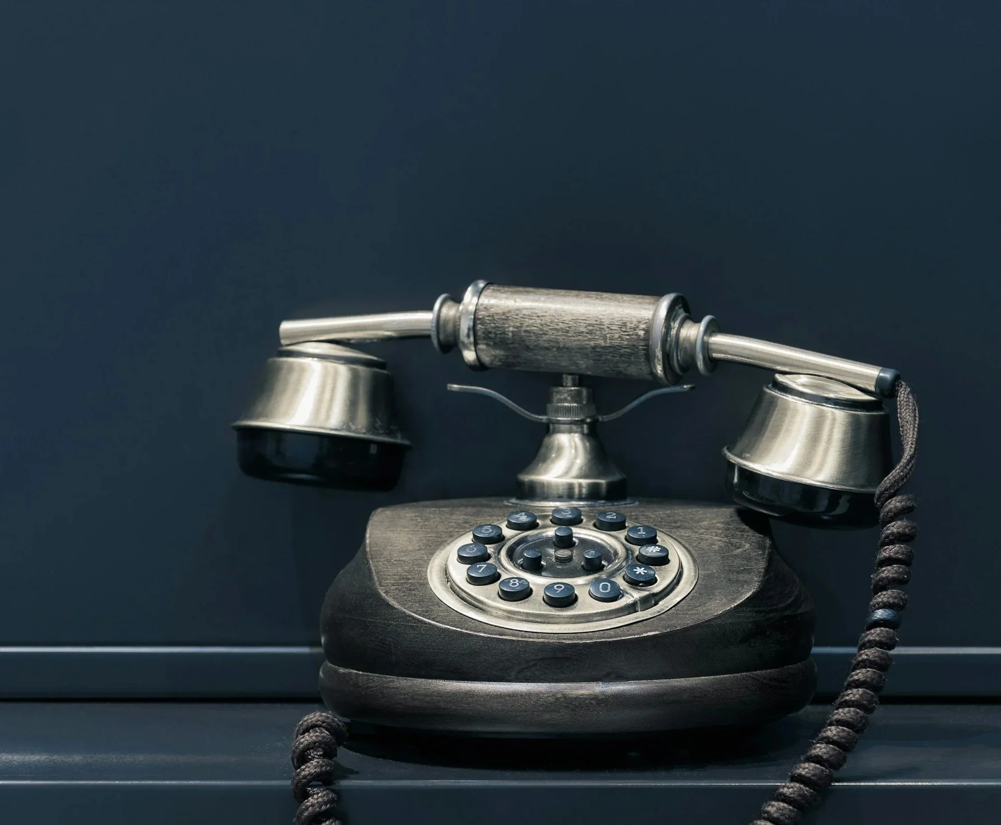 Vintage rotary telephone with a metal receiver, wooden handle, and black and metallic components, placed on a dark surface against a dark background.