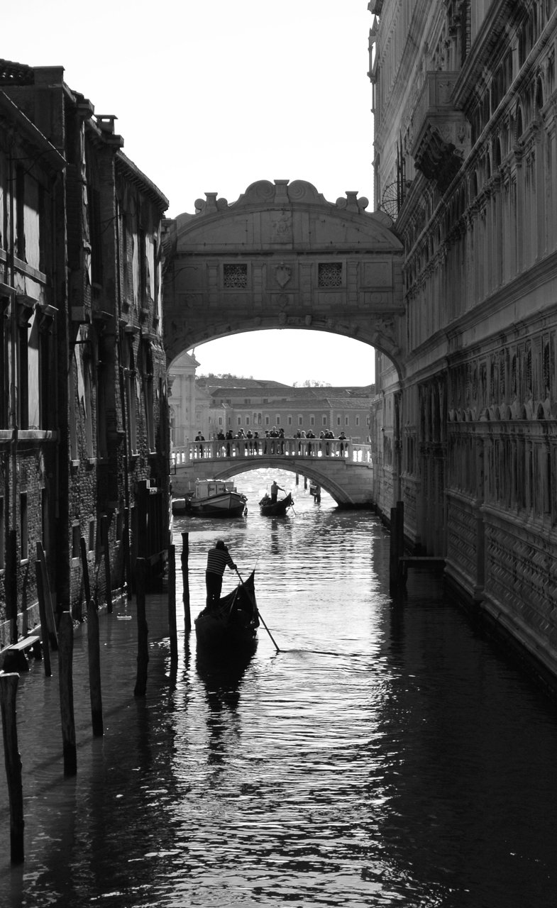 A black and white photograph of a canal in Venice, Italy, with a gondola and gondolier in the foreground, a bridge with pedestrians in the middle ground, and historic buildings lining the canal.