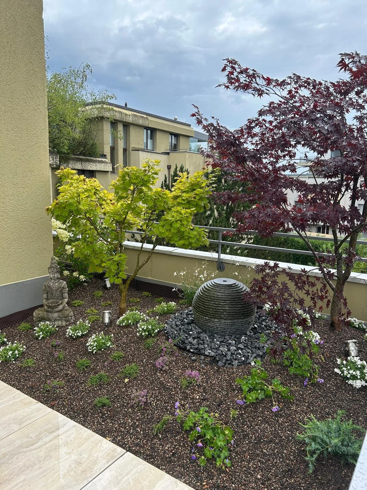 Balkongarten mit verschiedenen Pflanzen, einem kleinen Wasserspiel, einer Buddha-Statue und bunten Blumen, mit Stadtgebäude im Hintergrund.