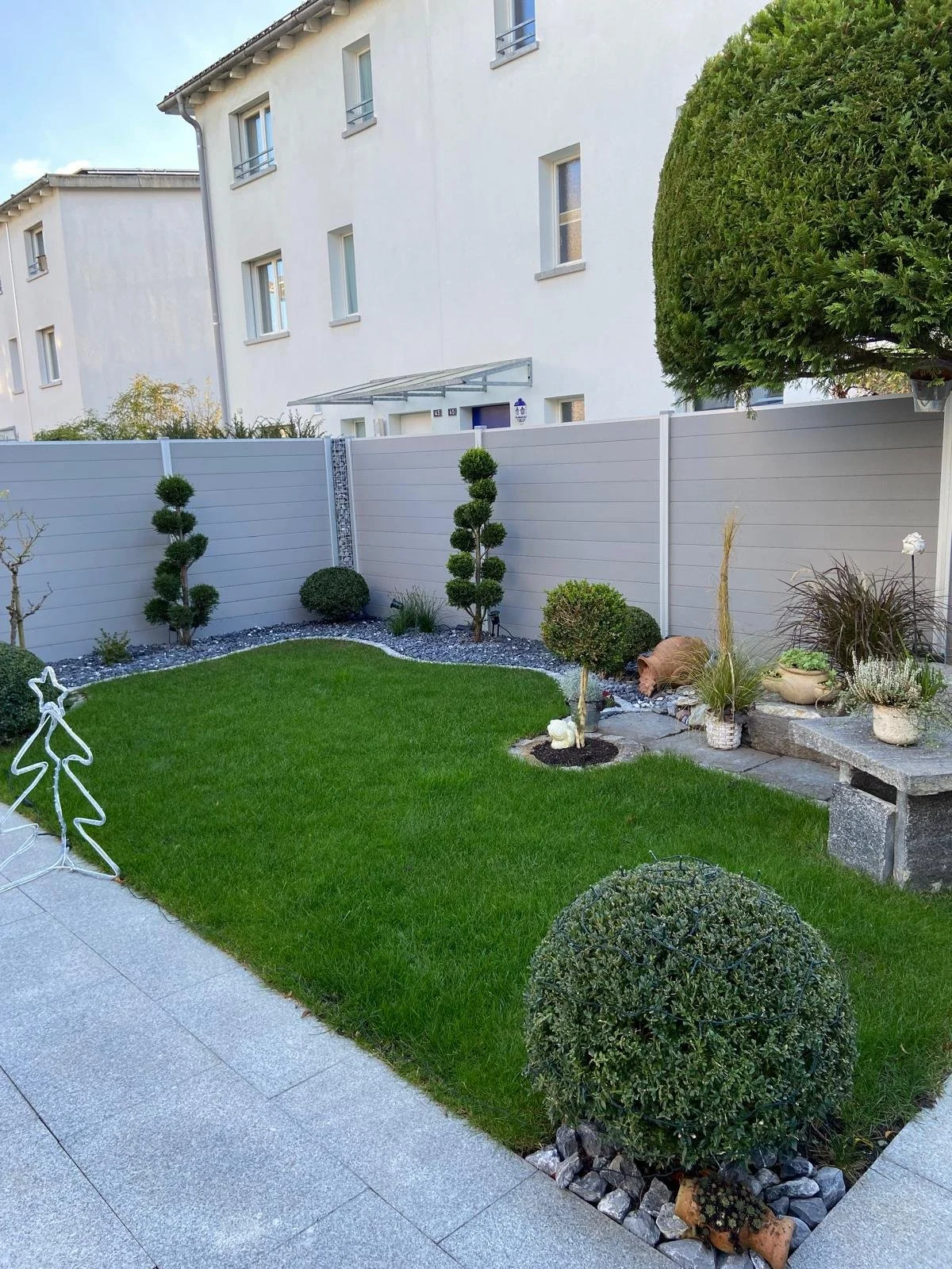 Ein gepflegter Garten mit grünem Rasen, ausgehöhlten Büschen, Topfpflanzen und einer weißen hölzernen Umzäunung. Im Hintergrund sind weiße Wohngebäude mit mehreren Fenstern sichtbar.