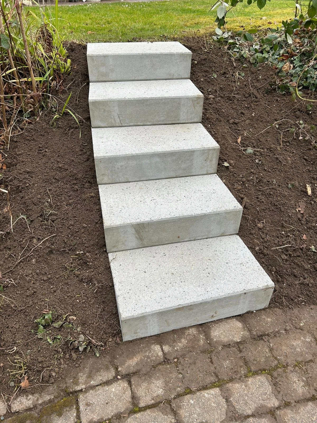 Vier Stufen aus grauem Stein in einem Garten, umgeben von Erde und Pflanzen, mit einer Rasenfläche im Hintergrund.
