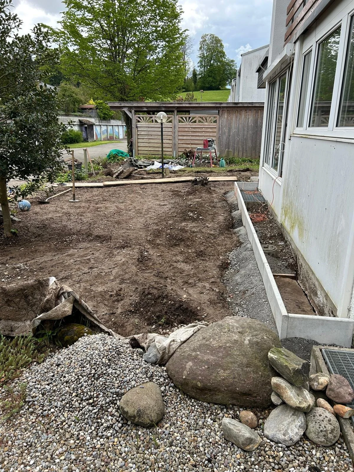 Ein Garten im Umbau mit Erde und Steinen, Bauarbeiten im Vordergrund, eine weiße Hauswand an der rechten Seite, Holzpflanzkübel, ein Scheinwerfer, eine Holzabsperrung im Hintergrund, grüne Bäume und ein bewölkter Himmel.