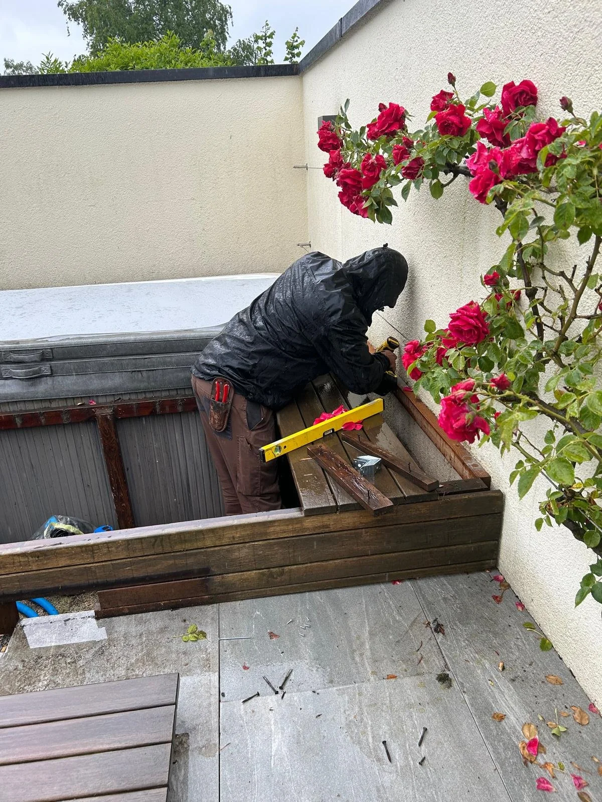 Eine Person trägt eine schwarze Regenjacke und arbeitet im Freien an einer Holzbank, die gegen eine weiße Wand gelehnt ist, bei Regen. Die Person benutzt ein Werkzeug, während um sie herum Werkzeuge und Schrauben auf dem Boden liegen. Eine Blume mit 