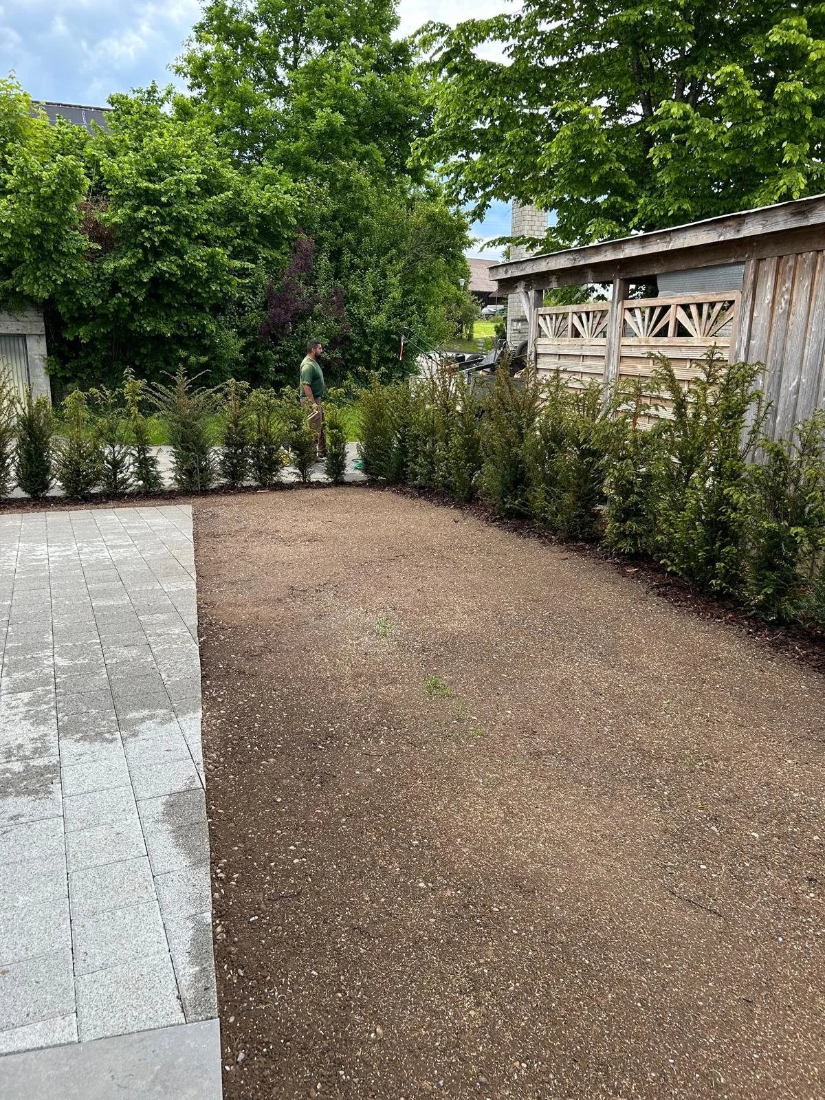 Garten mit Baum, Hecke und Holzpavillon, im Hintergrund ein Mann beim Gartenarbeiten, auf einem großen gefliesten Bereich ist der Boden frisch gestaltet.