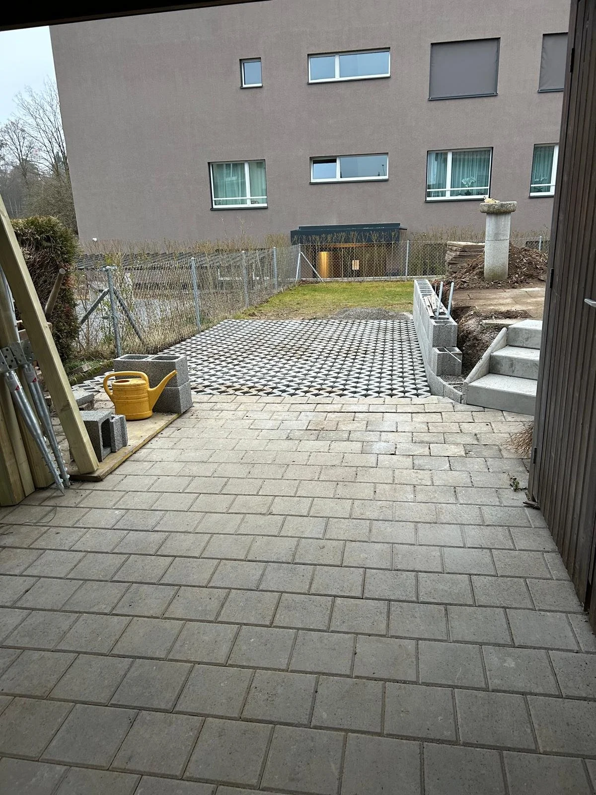 Ein Gartenbereich mit Pflastersteinen, einer grassbewachsenen Fläche, einer Mauer mit Zäunen, einer Terrasse mit Treppe und einem Wasserbehälter, während im Hintergrund ein Mehrfamilienhaus sichtbar ist.