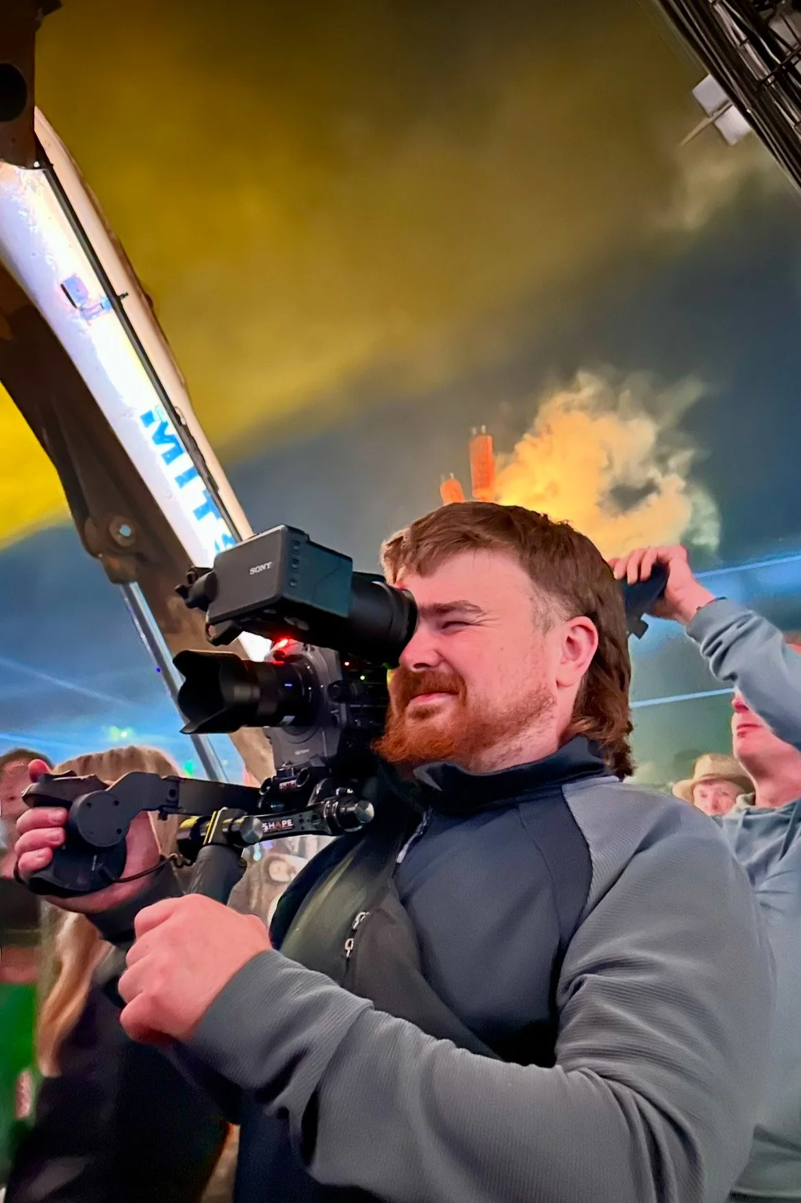 A man with a beard and long hair using a professional video camera, aiming through the viewfinder, with a fire and smoke in the background.