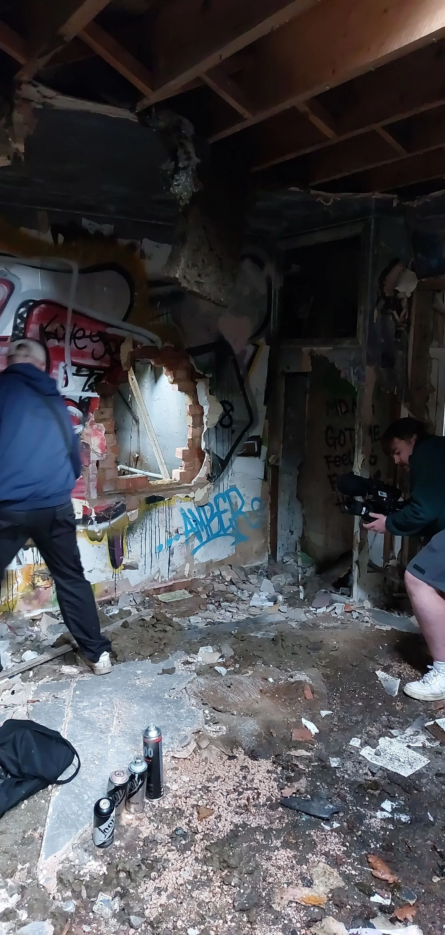 Abandoned and vandalized building interior with exposed wooden ceiling beams, graffiti on the wall, rubble on the floor, and two people inspecting the space.