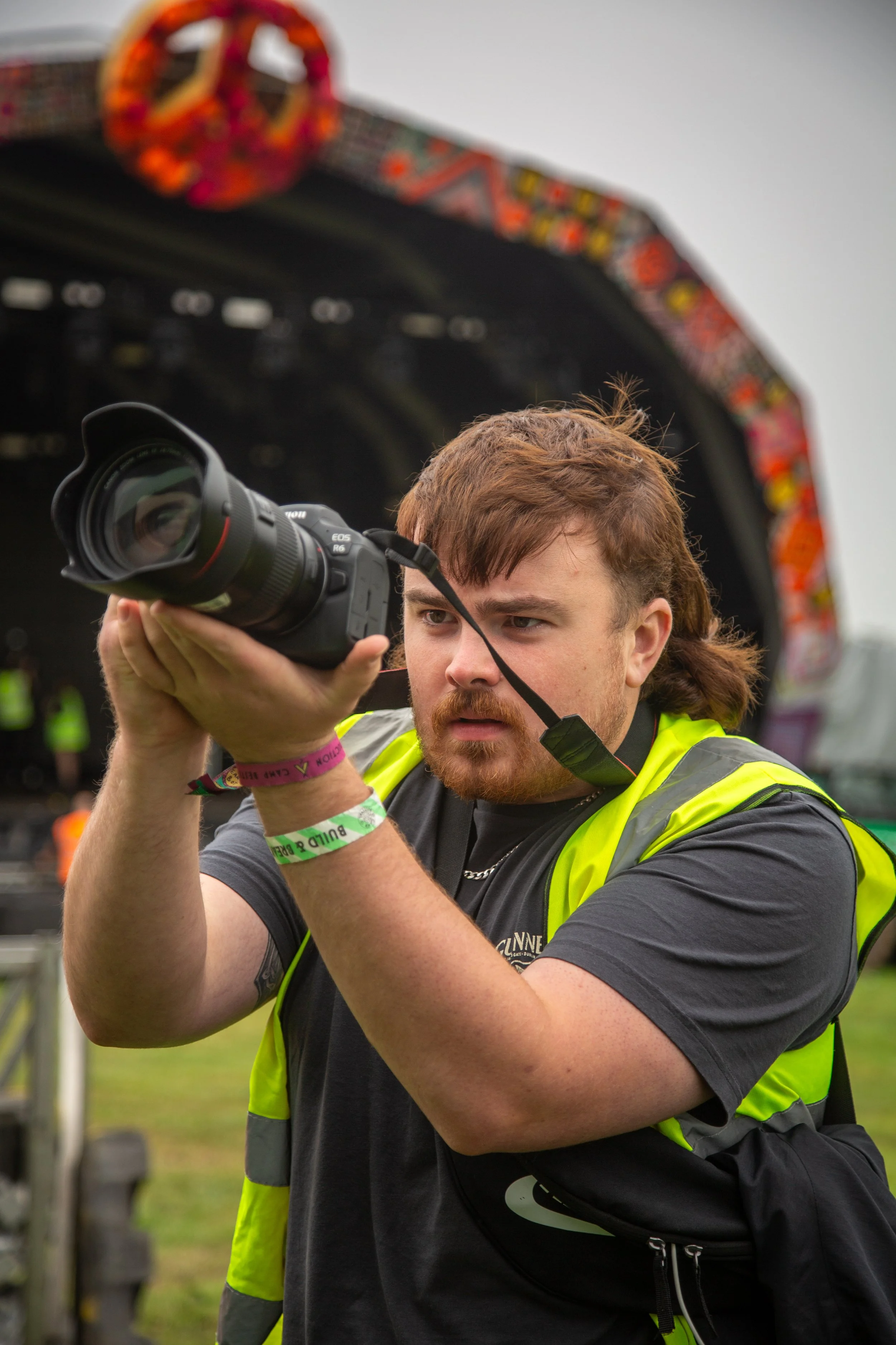 A man with a beard and brown hair wearing a high-visibility vest is looking through the lens of a professional camera at an outdoor event.