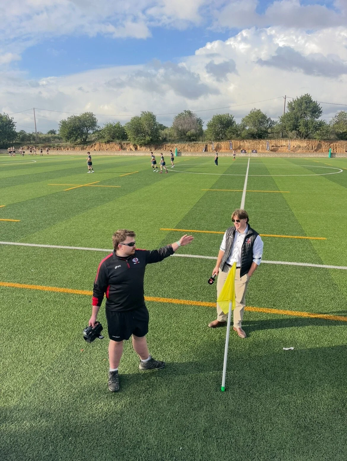 Two men standing on a football field, one gesturing towards the distance, another holding a camera near a corner flag. In the background, players practicing on the field, with grassy hill and trees behind.
