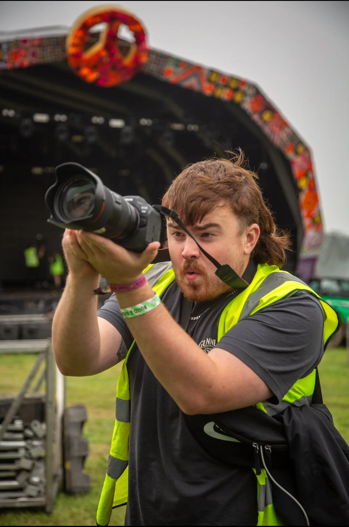 A man with reddish-brown hair and a beard wearing a black t-shirt, a yellow safety vest, and wristbands, holding a camera up to his face at an outdoor music festival with a stage and colorful decorations in the background.
